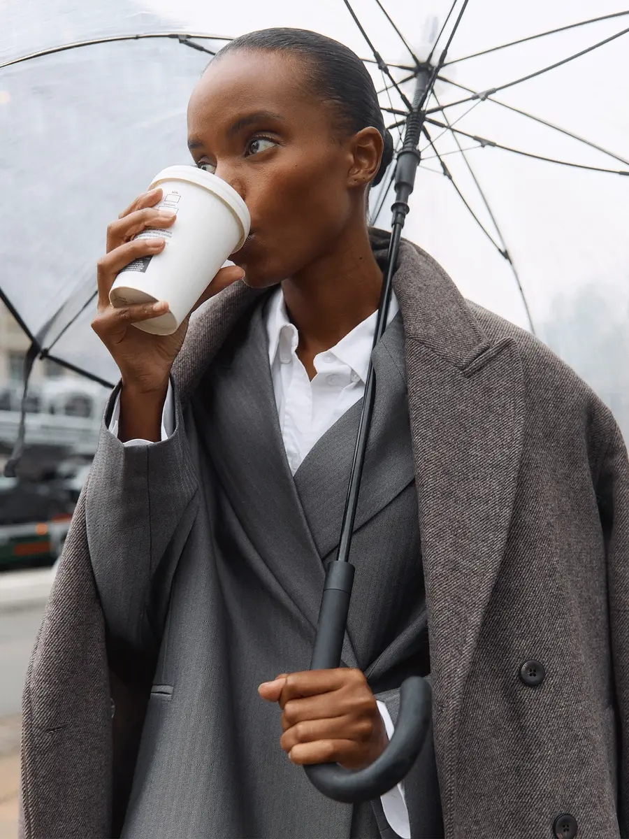Elegante vrouw in zakelijke kleding met koffie en transparante paraplu op straat.