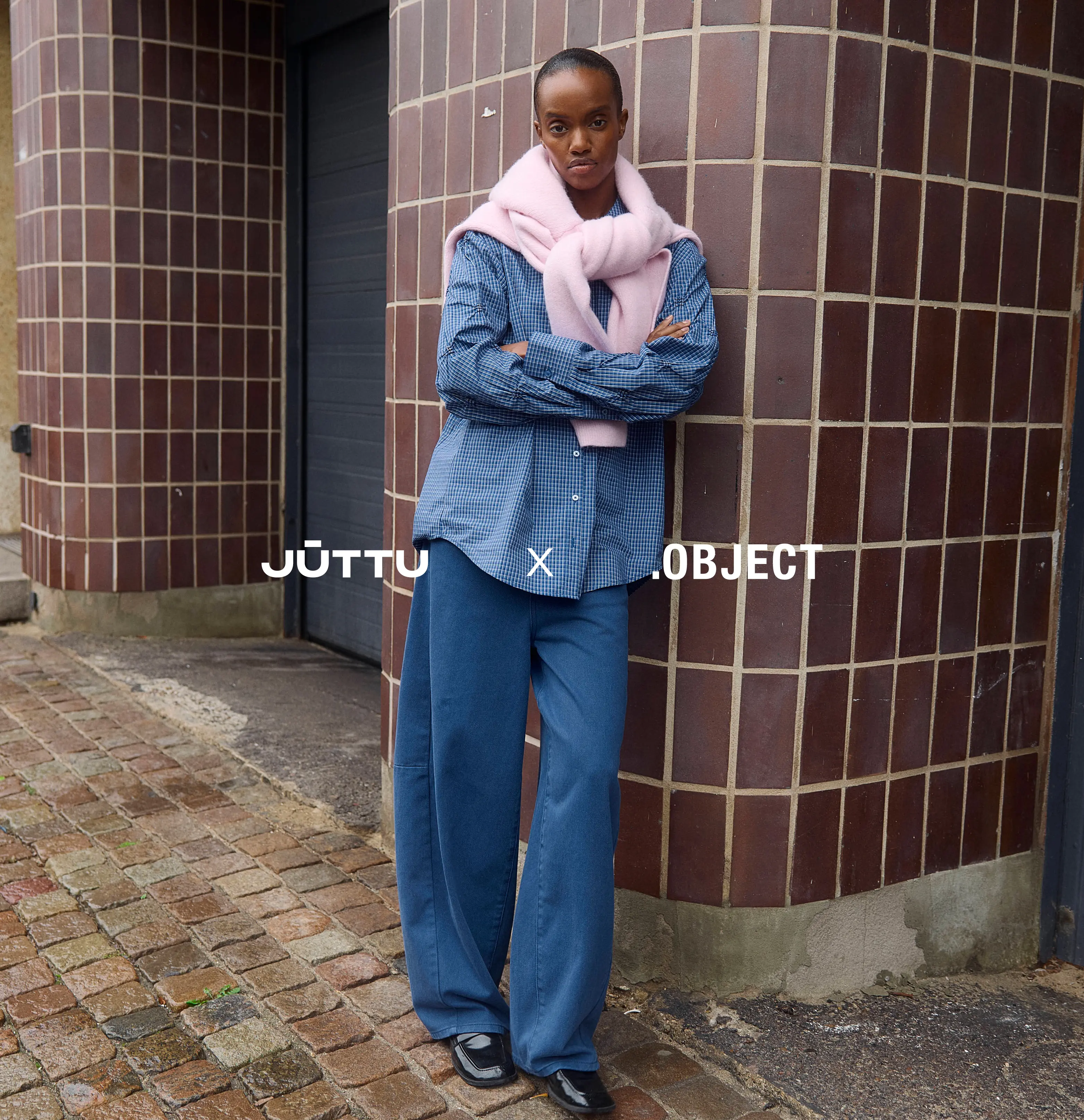 Short-haired woman in pink sweater and blue wide-leg pants stands by a brown tiled wall.