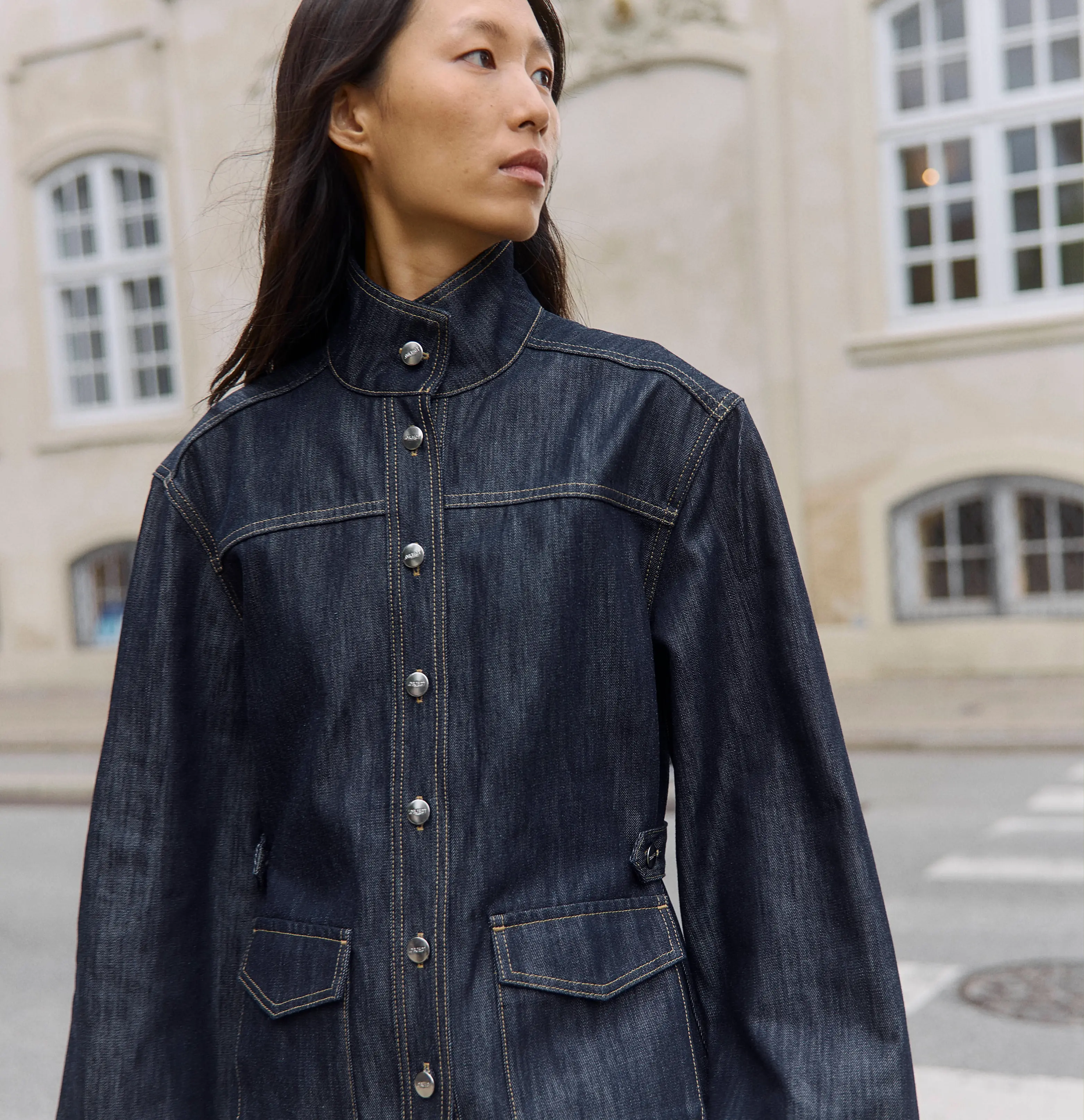 Asian woman in a dark denim jacket looking away against an urban building.
