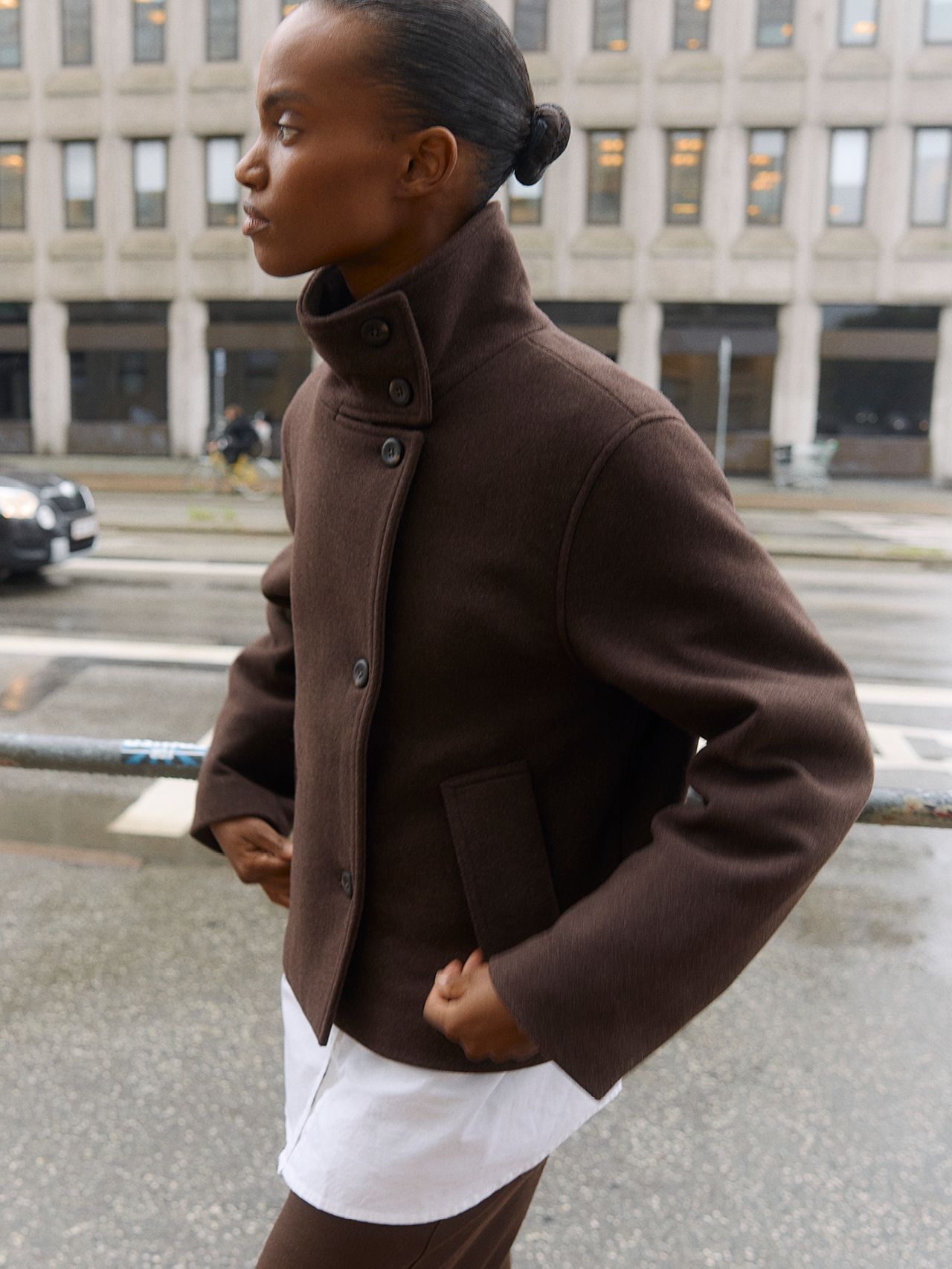 Woman in a brown jacket and pants, hands in pockets, walking on a city street.