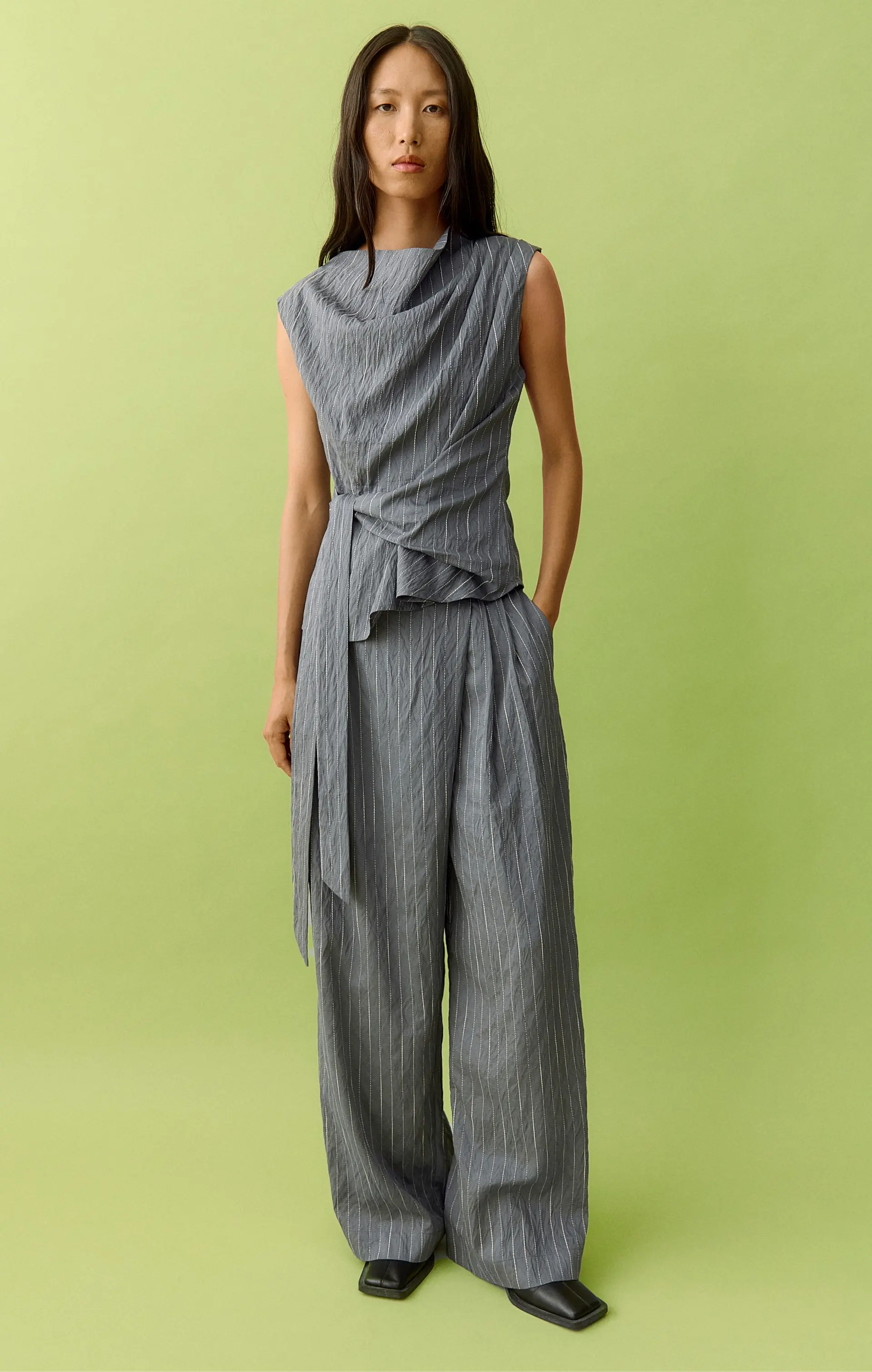 Woman in grey pinstripe draped top and wide-leg pants against a lime green background