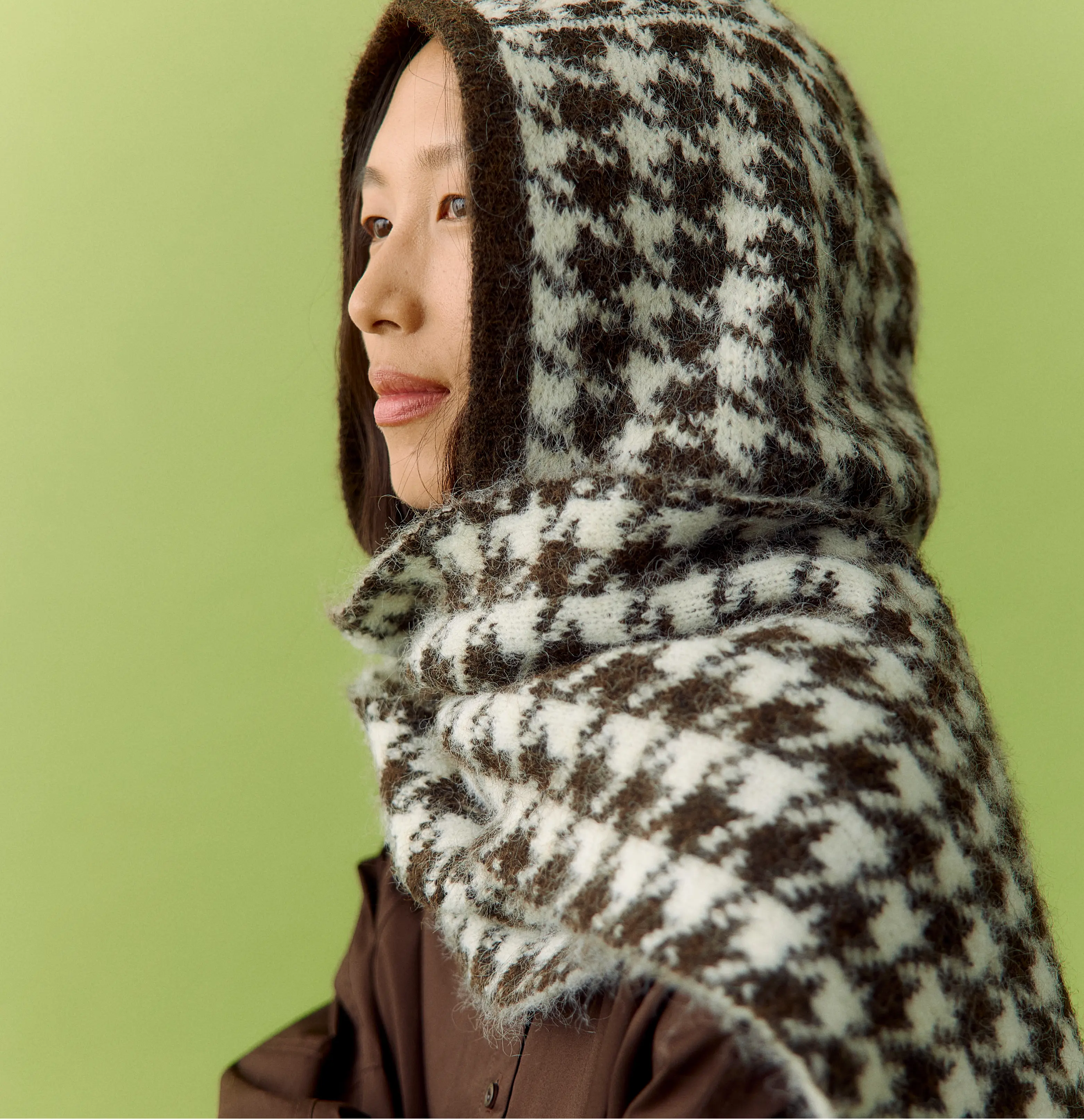 Asian woman in profile wearing a brown houndstooth hood and knitted scarf on green background