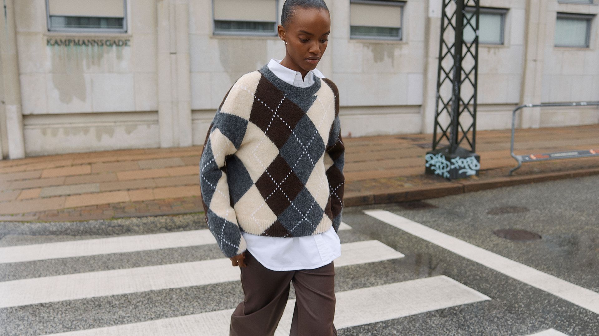 Femme en pull argyle oversize et pantalon marron traversant Kampmannsgade.