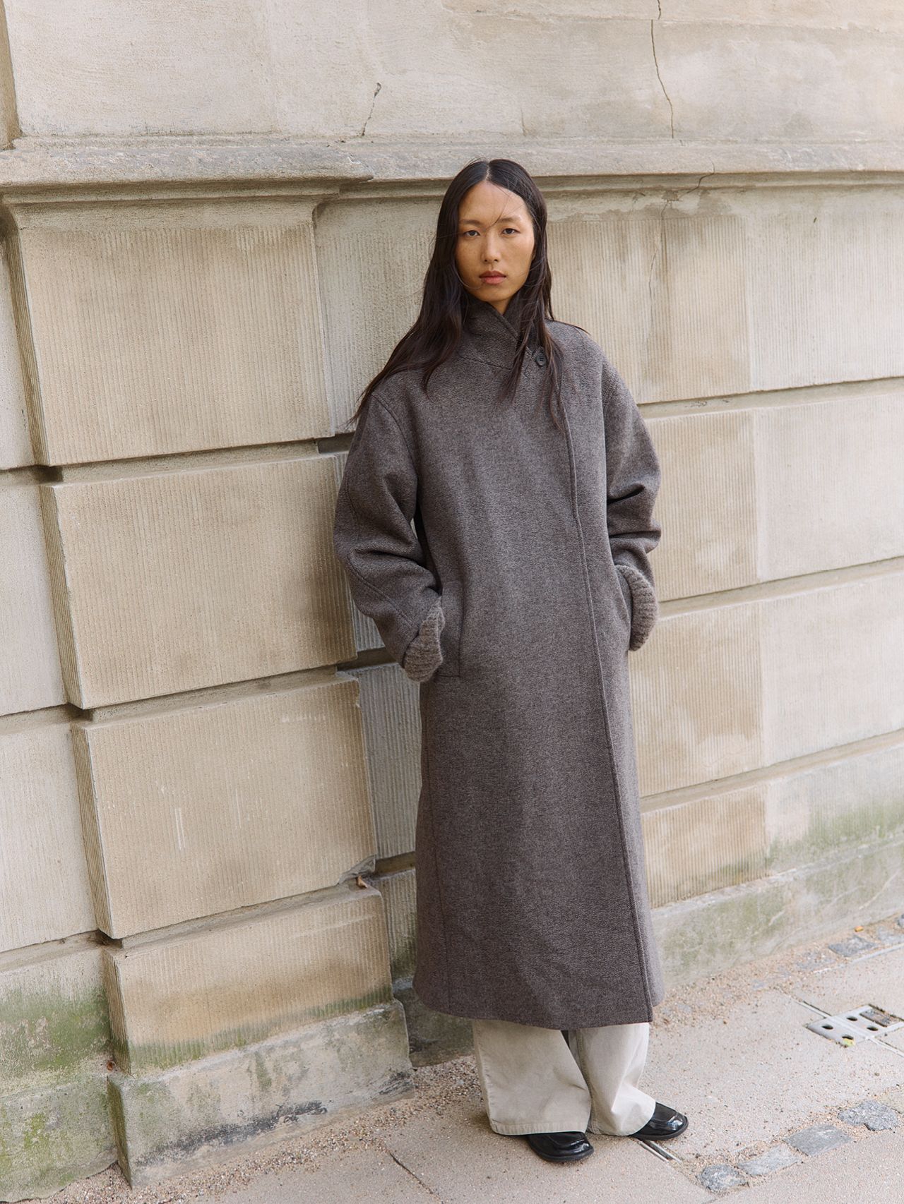 Asian fashion model in a long wool coat, wide-leg pants and loafers by a stone wall
