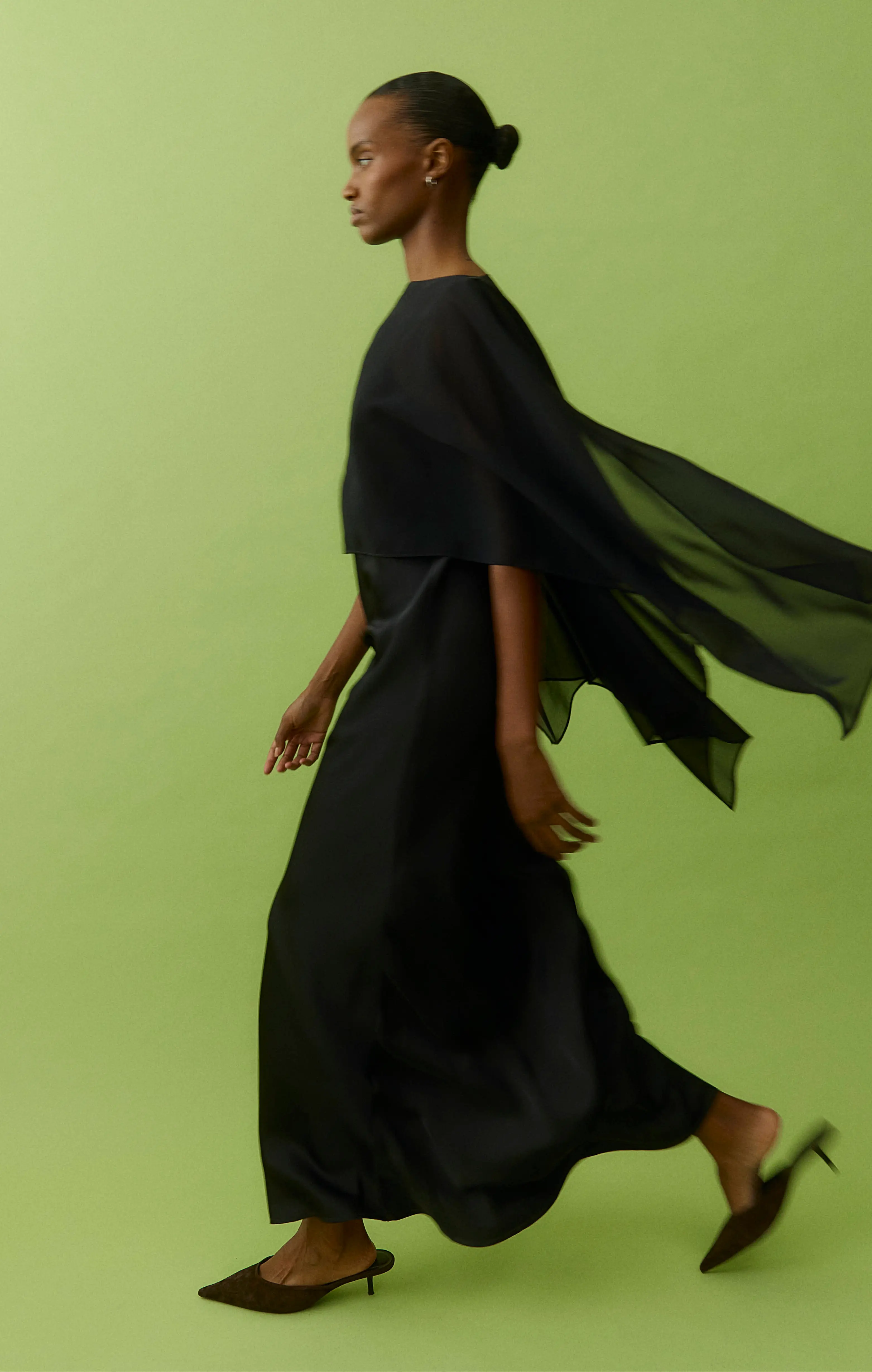Woman in a flowing black maxi dress and heels walking against a lime green background
