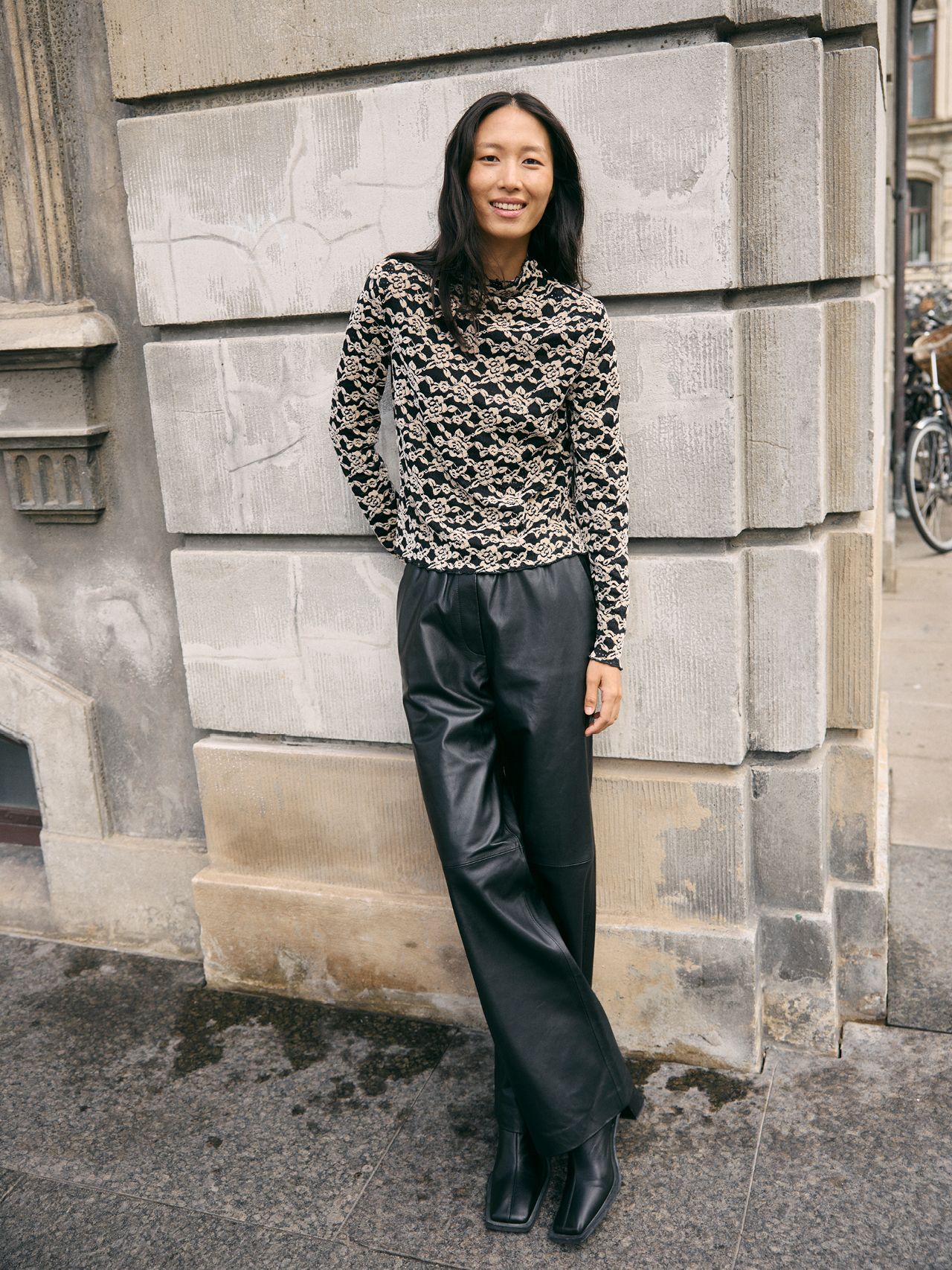 Smiling Asian woman in a patterned top, black leather pants and boots by a stone building