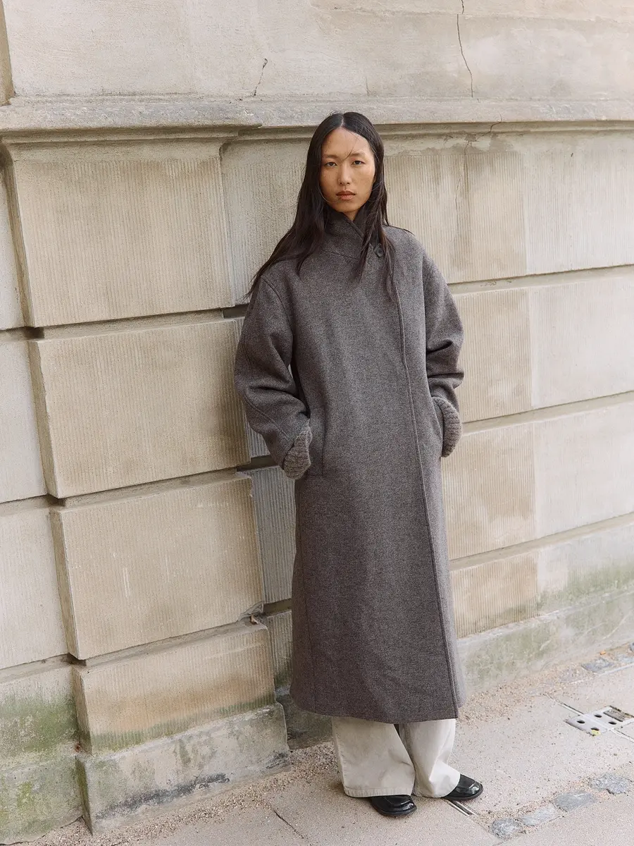 Woman in oversized gray wool coat and light wide-leg pants by a stone wall.