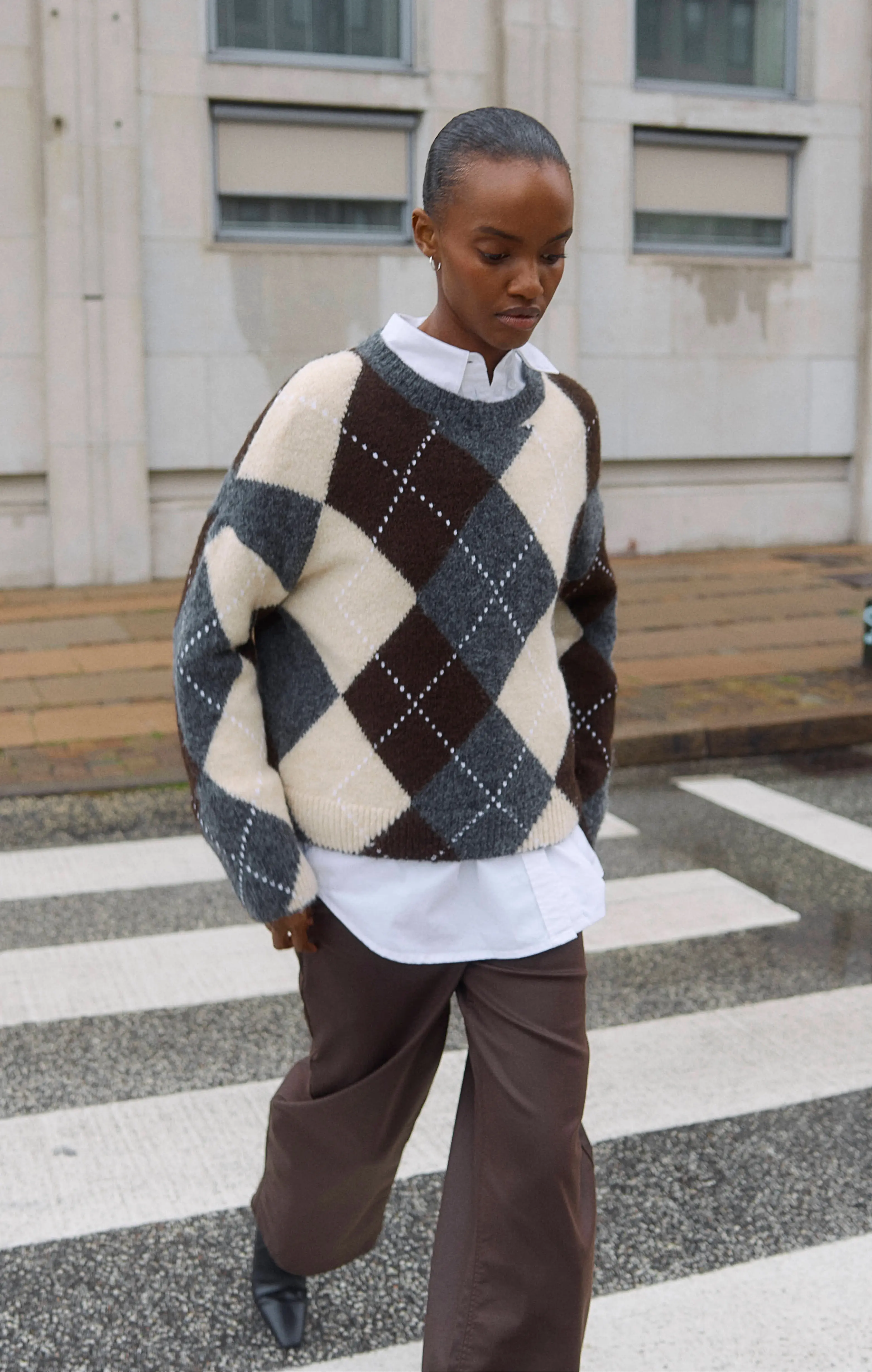 Woman in argyle sweater and brown pants walking across an urban crosswalk.