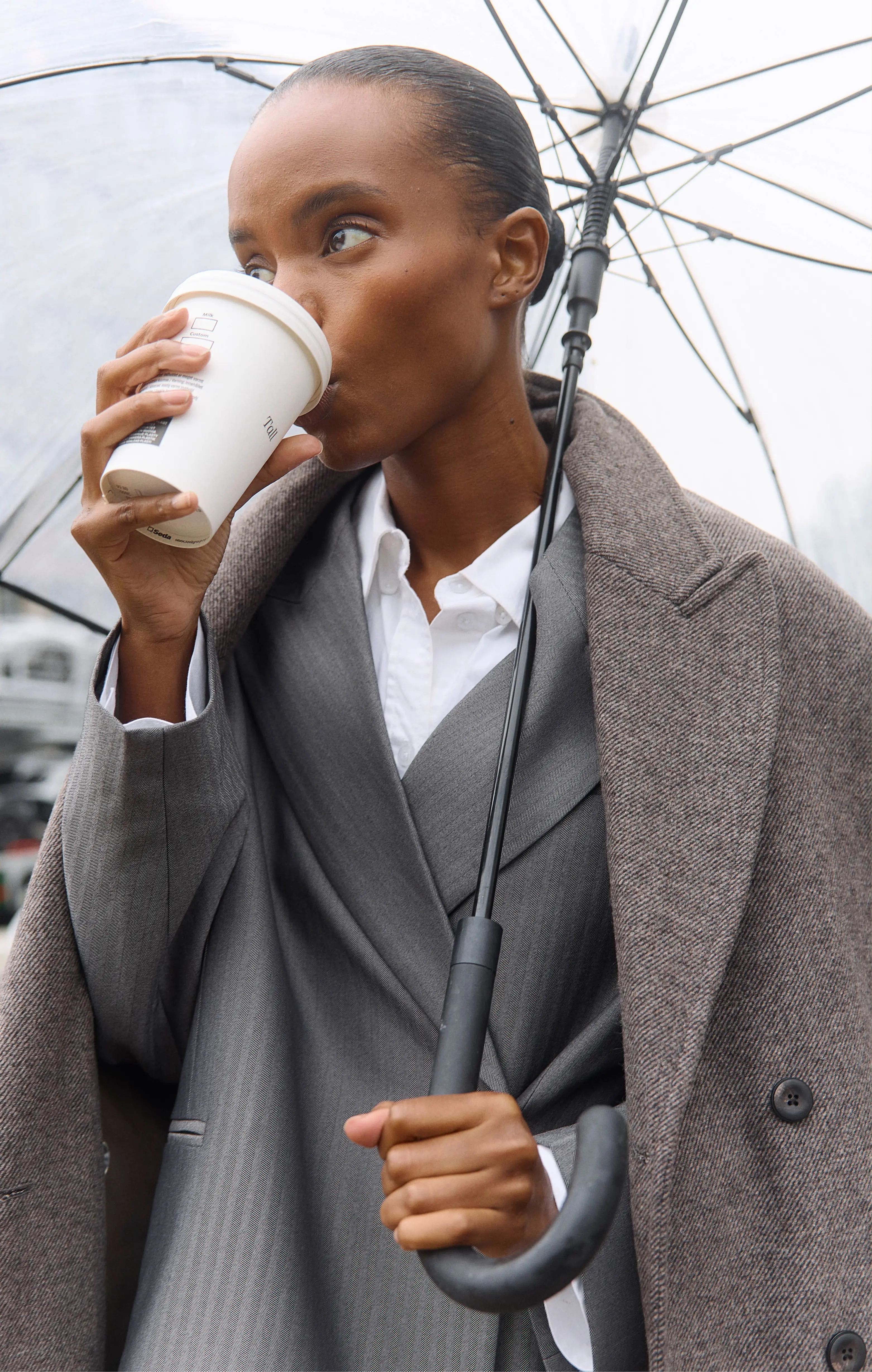Dark-skinned woman in a brown coat holding an umbrella and drinking coffee in the rain