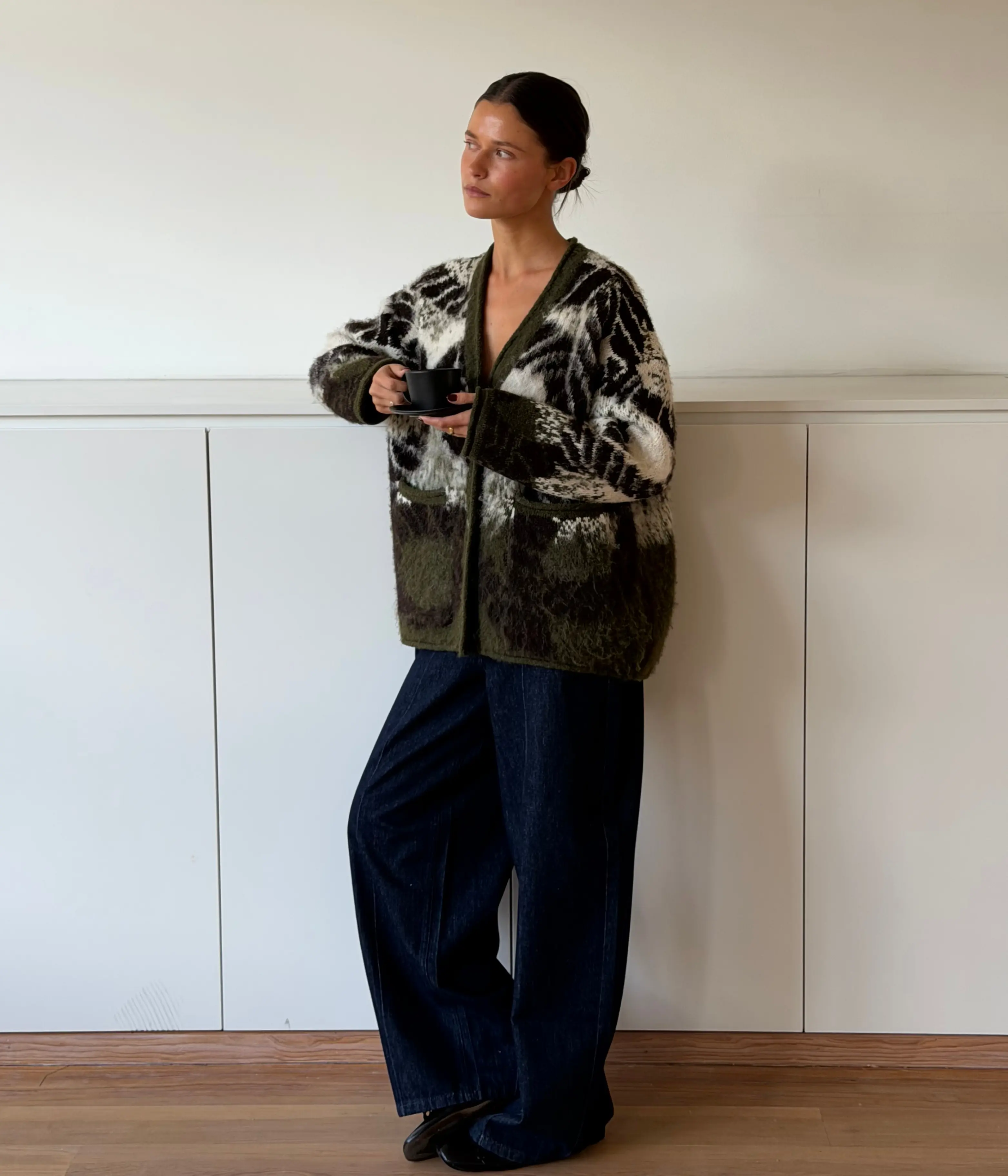 Brunette vrouw in oversized gebreid vest en wijde jeans leunend binnen met koffiebeker
