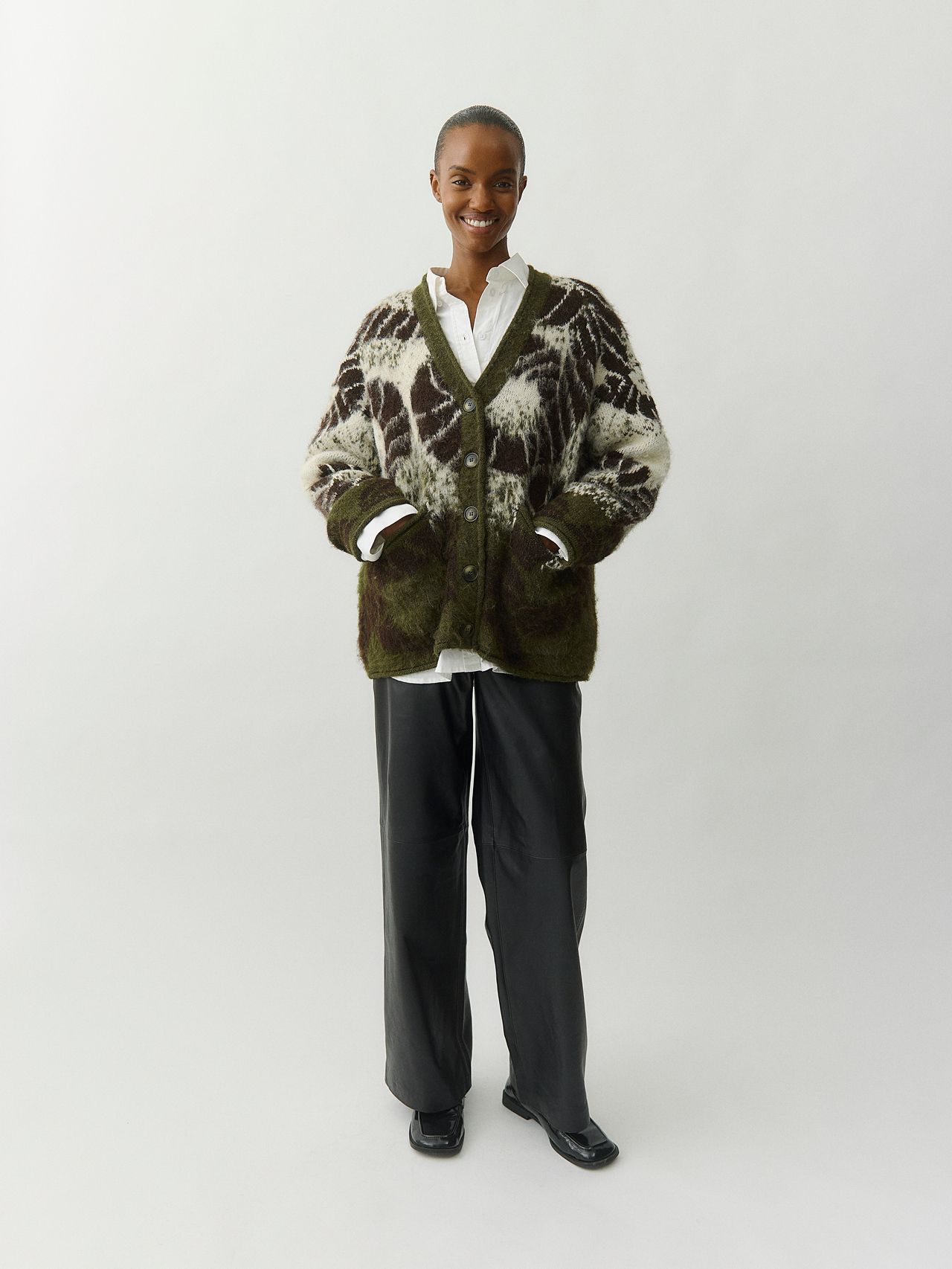 Smiling model in a green mohair cardigan, white shirt, black leather pants and loafers.