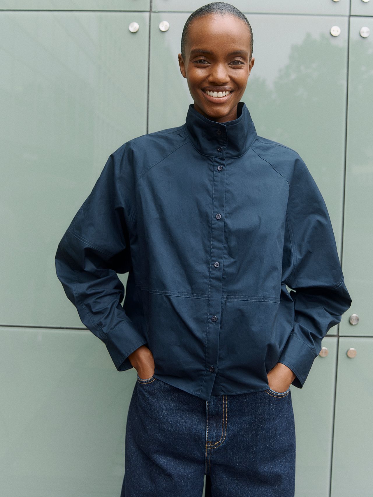 Smilende kvinne med kort hår, marineblå skjorte og mørke jeans foran grønn vegg