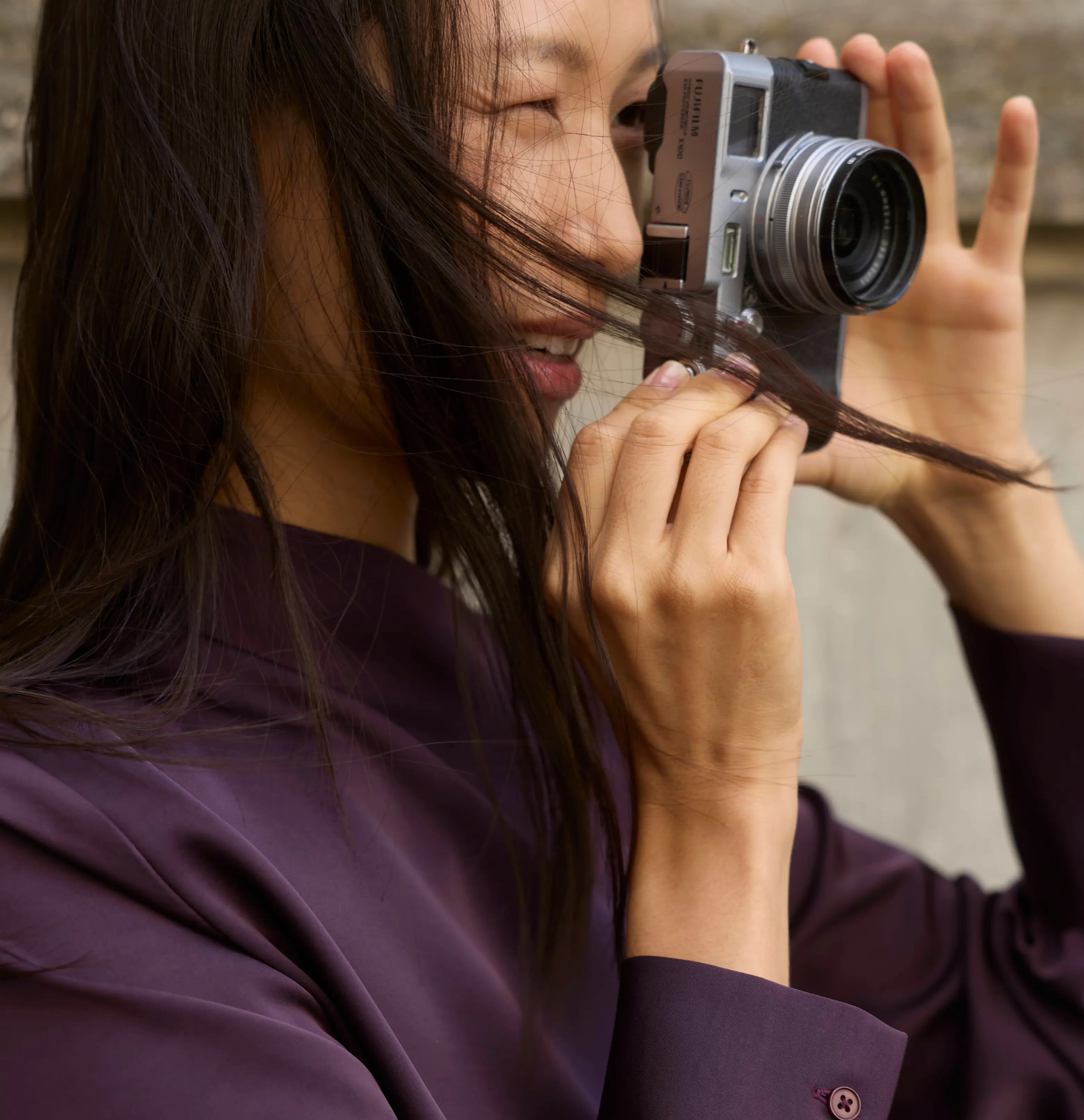 Smiling Asian woman with a Fujifilm camera, her long dark hair blowing outdoors.