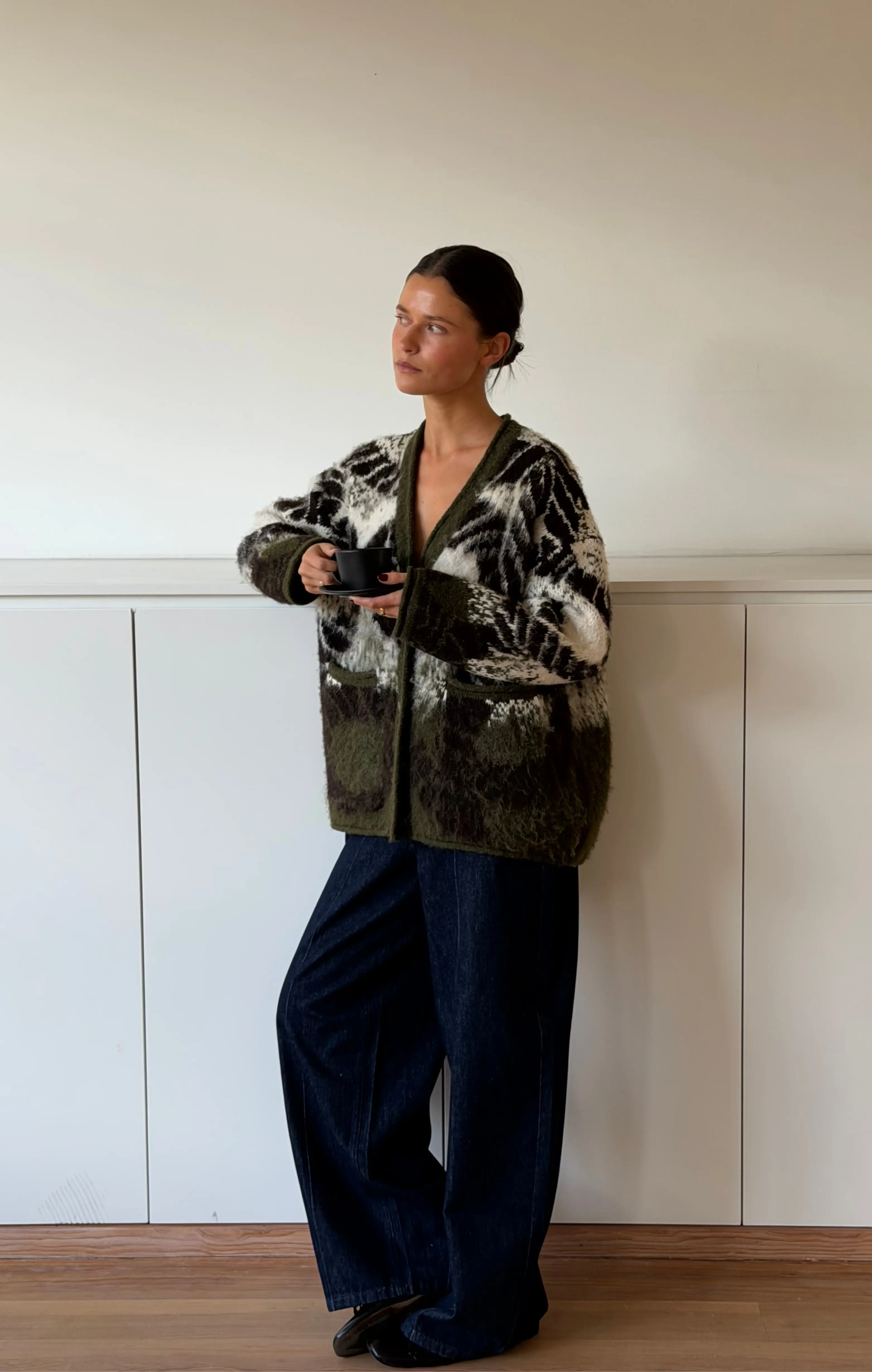 Vrouw in groene shaggy cardigan met wijde spijkerbroek, kopje in hand, kijkt opzij indoors