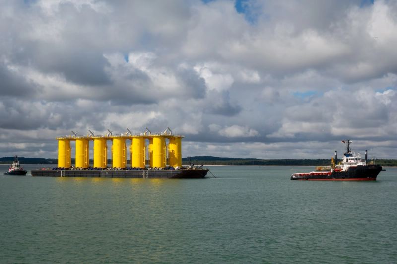 Offshore wind foundations under tow on a barge with a tug alongside
