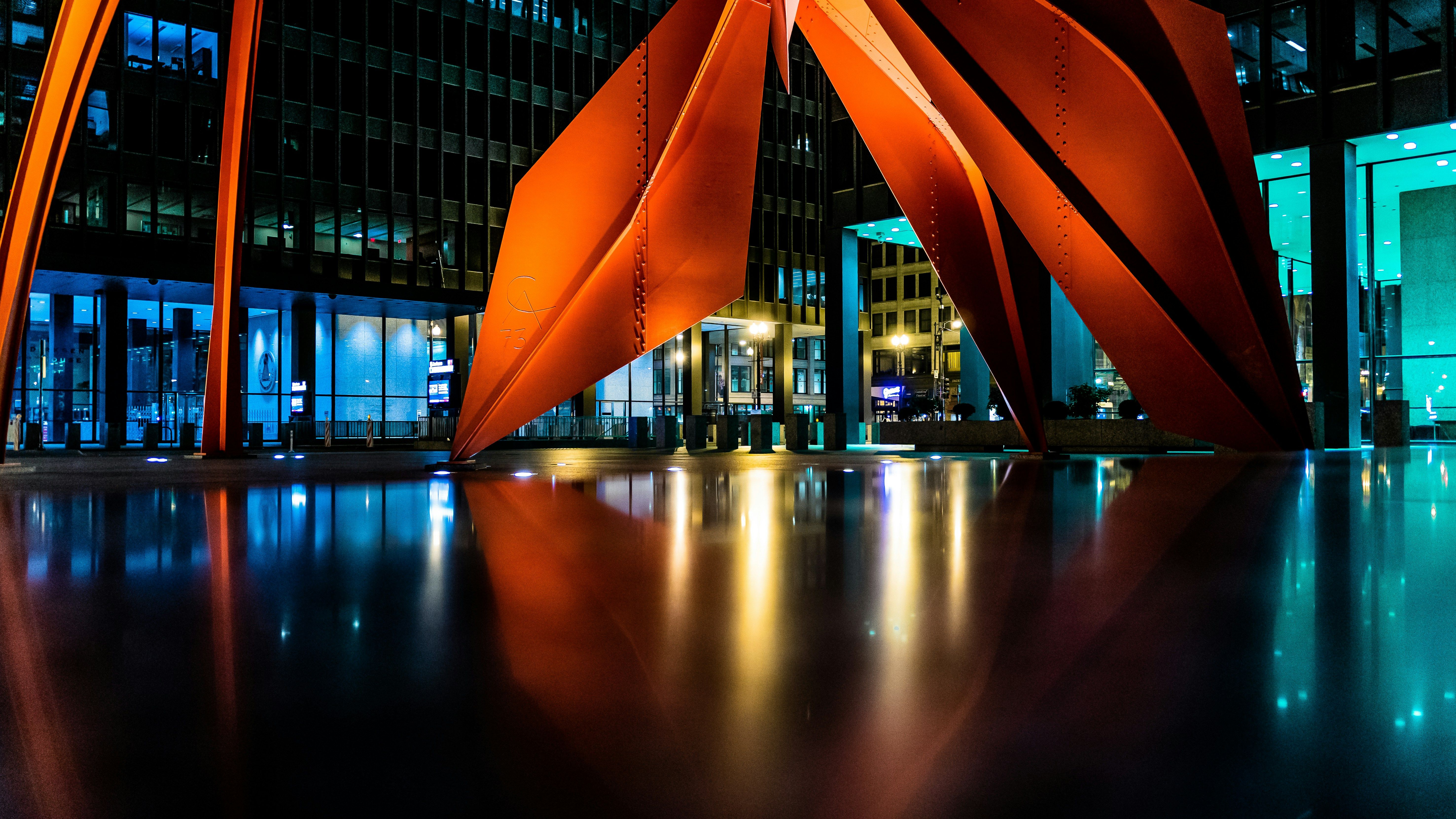 A night color photograph of the red Flamingo steel sculpture downtown Chicago at night, with shades of soft teal and blue lighting coming from the neighboring building lobbies. 