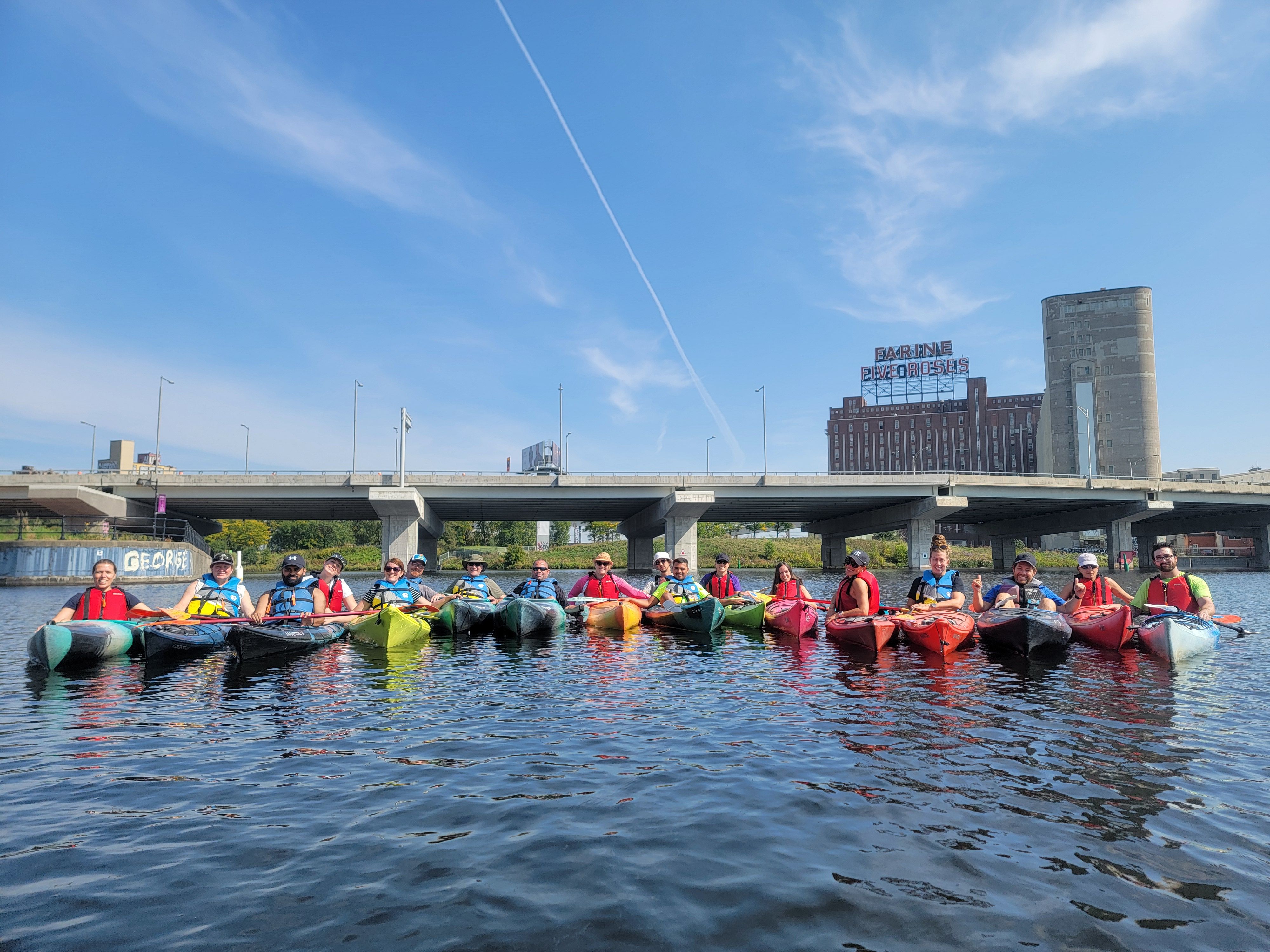 A group of kayakers paddling on the Lachine Canal in front of the illuminated Farine Five Roses neon sign in Montreal.