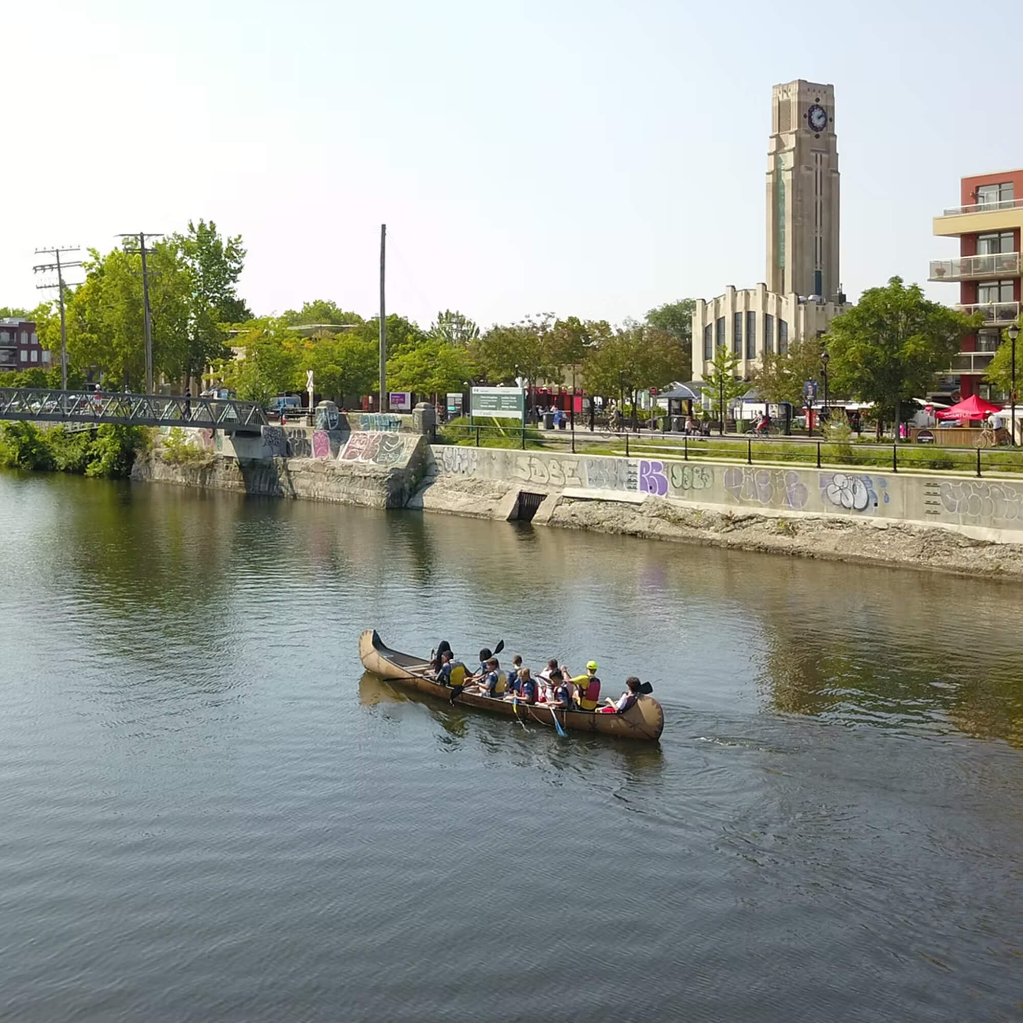 A group paddling a large rabaska canoe along the Lachine Canal past the iconic Atwater Market building in Montreal.