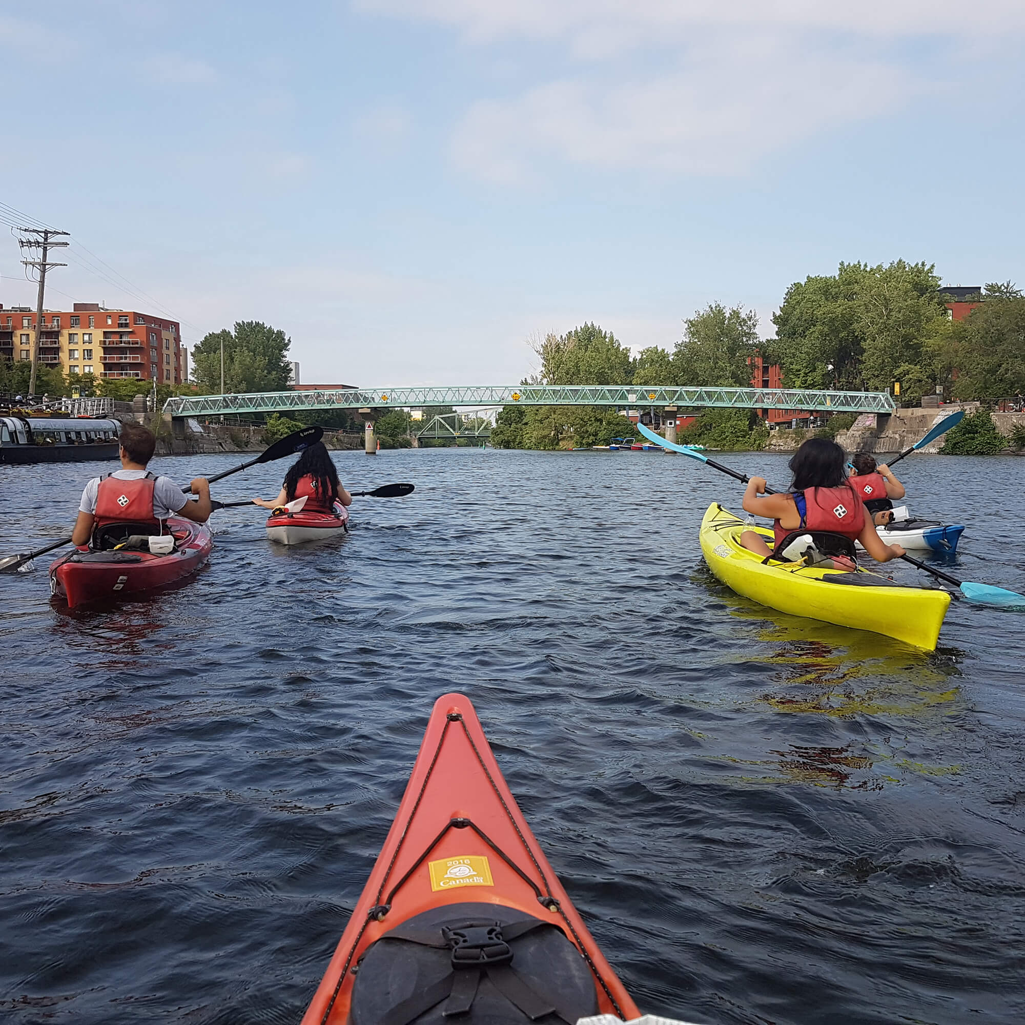 A first-person view from a kayak paddling with a group under the Atwater footbridge along the Lachine Canal in Montreal.