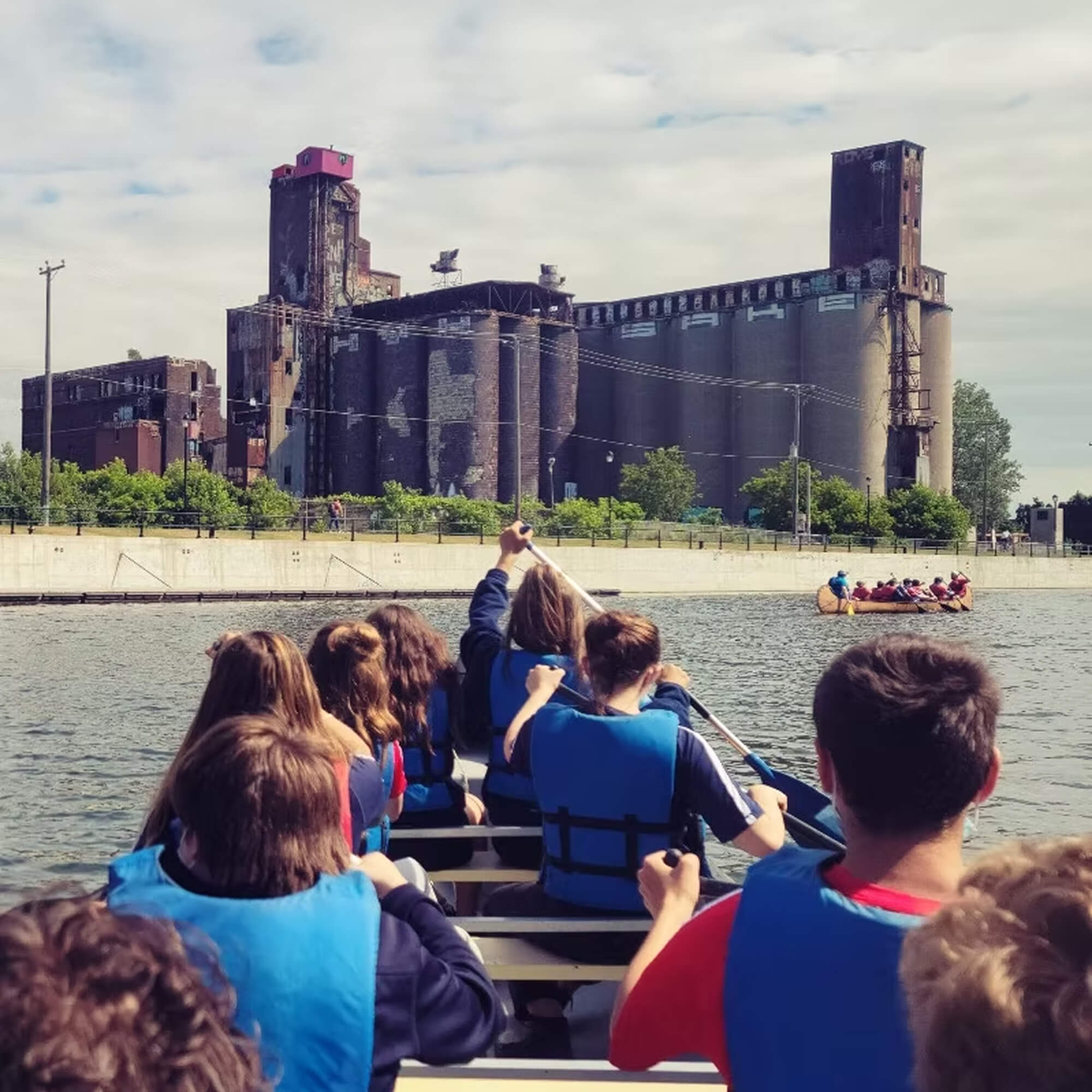 A first-person view from a boat showing a group of kids wearing blue lifejackets paddling down the Lachine Canal in Montreal towards the iconic, abandoned Canada Malting silos with the famous pink cottage perched on top.