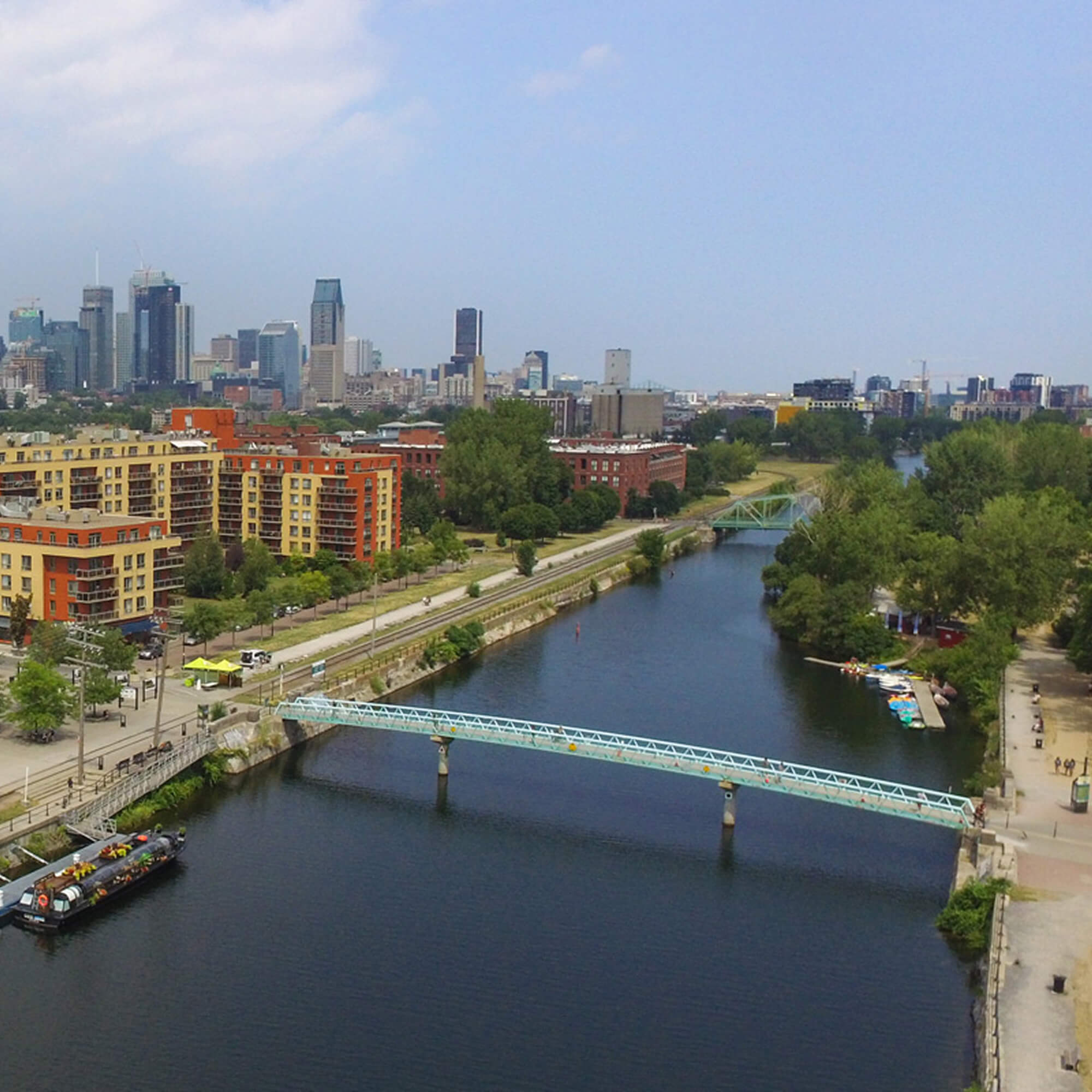 An aerial view of the Lachine Canal in Montreal showing pedestrian footbridges, the surrounding neighborhood, and the downtown skyline in the background.