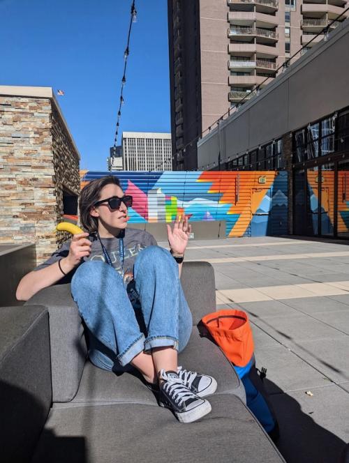 Brunette with jeans sitting on a sofa with an orange and blue backpack against a multicolor mural. She is holding a banana and speaking.