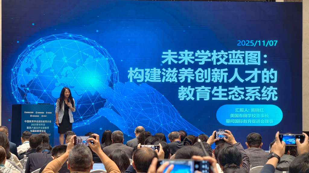 Woman in a blazer speaking at a podium with a large digital globe and Chinese text on a blue screen behind her, while an audience records the presentation with phones.