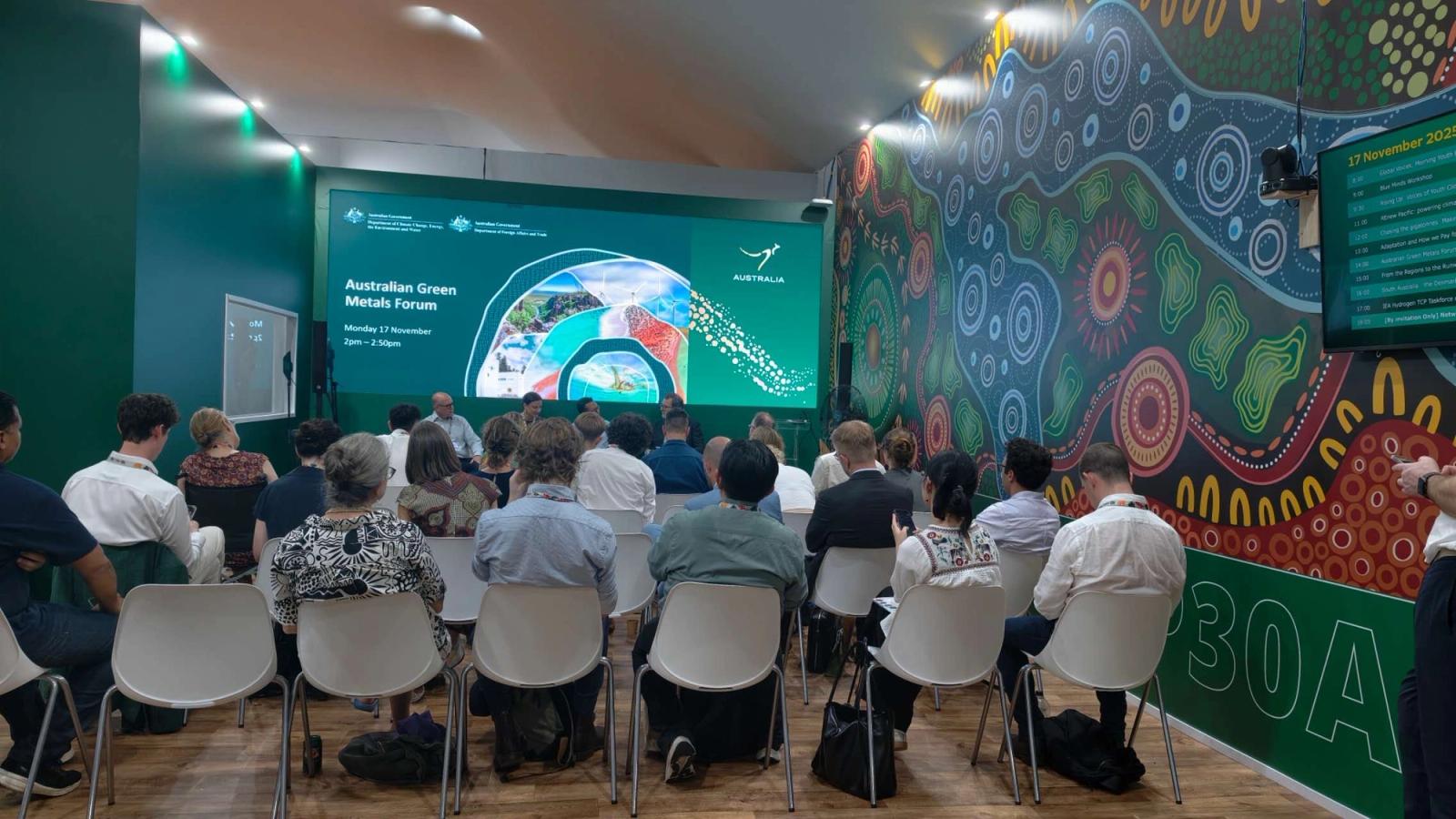 Audience attending the Australian Green Metals Forum seated in a room with colorful Indigenous-style wall art and a large screen displaying the event information.