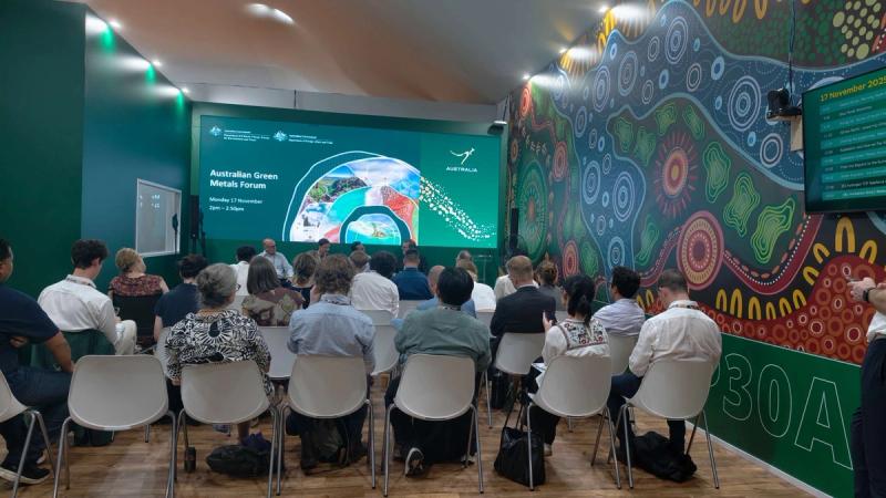 Audience attending the Australian Green Metals Forum seated in a room with colorful Indigenous-style wall art and a large screen displaying the event information.