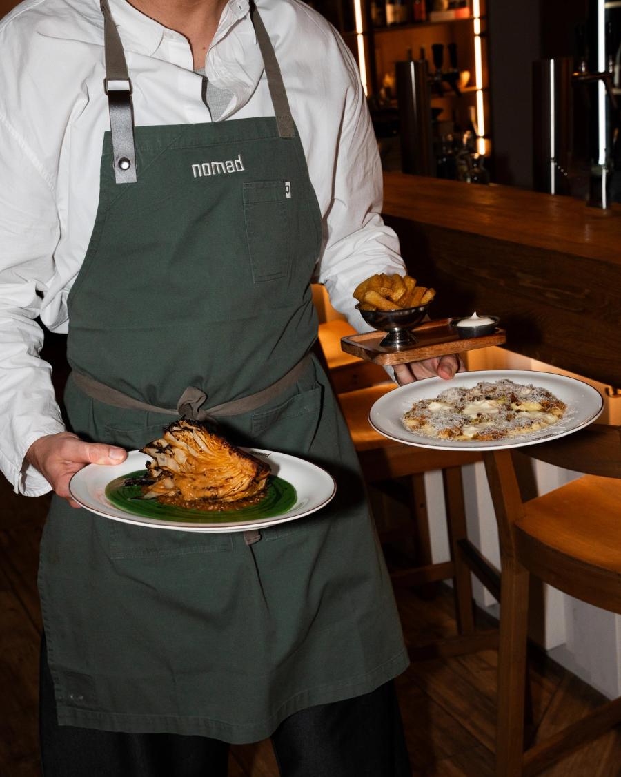 Server in a green Nomad apron bringing grilled pointed cabbage, handmade pasta and fries to the table