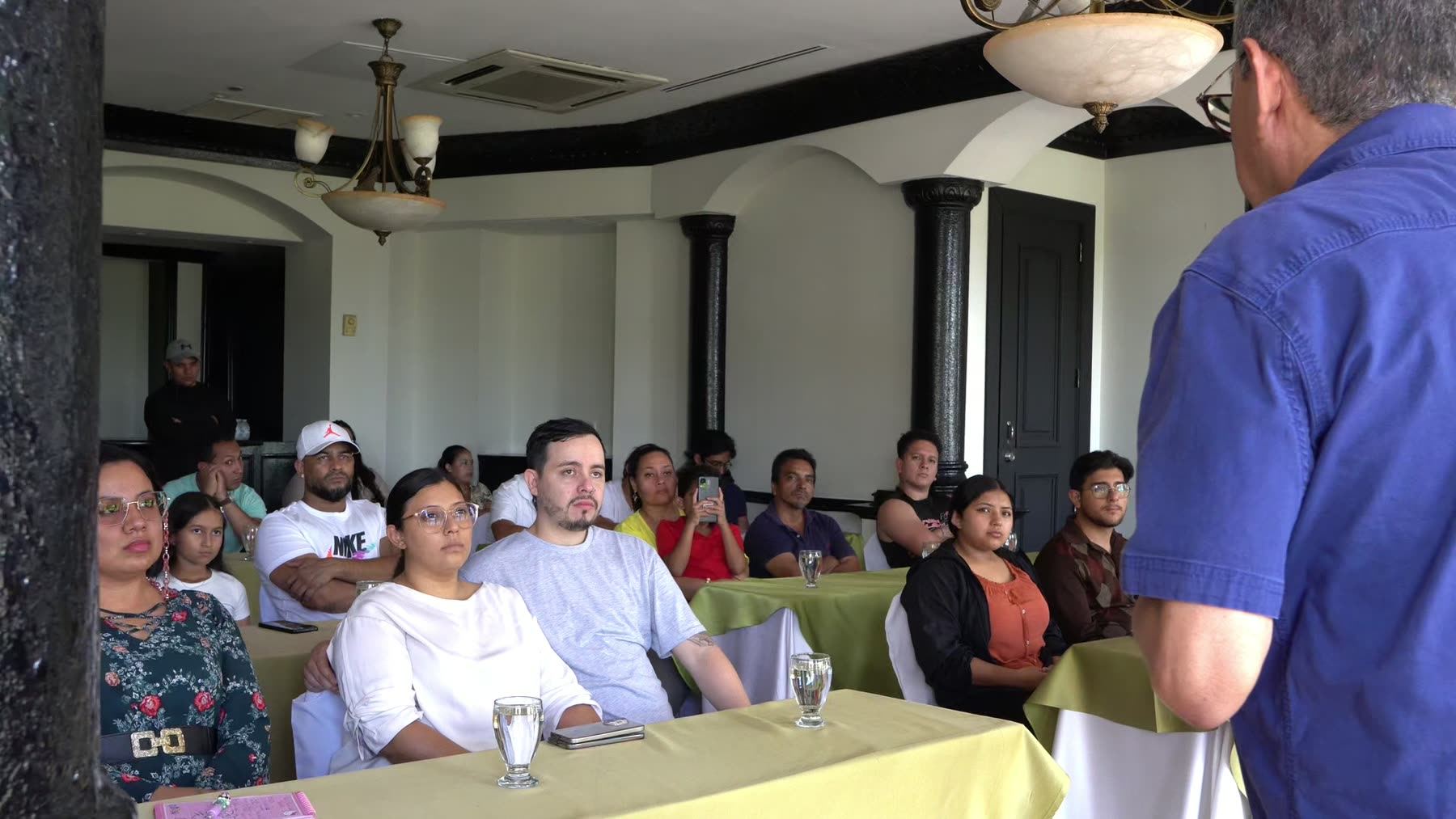 Un salón con las familias sentadas en mesas, frente a una pantalla, mientras un expositor presenta el proyecto.