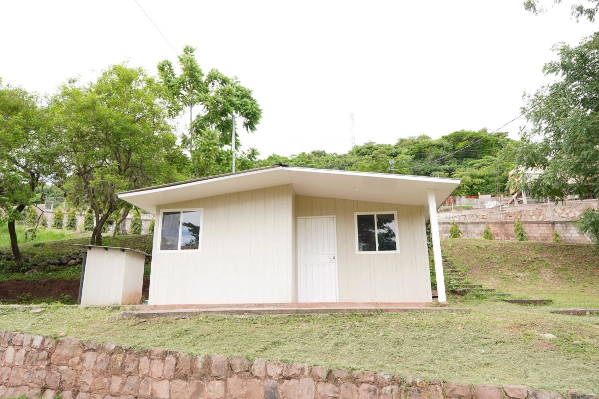 La casa modelo vista de lado, con su corredor cubierto y el muro de piedra al frente.