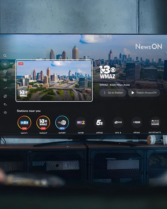 Back view of two persons sitting on a bench, watching their smart TV that is displaying the home screen of the NewsON connected TV app.