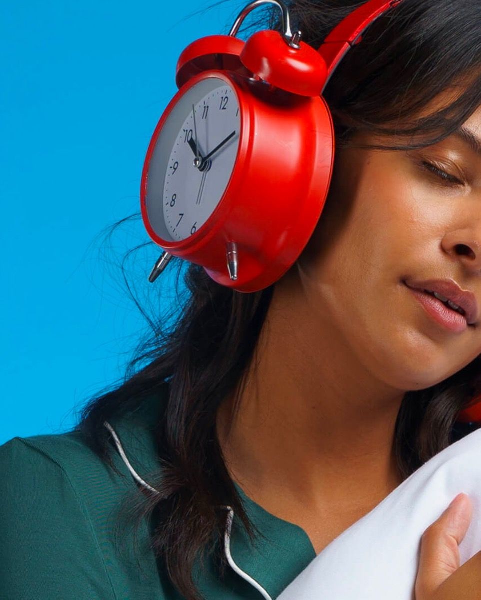 A woman with her eyes closed holding a pillow. Her over-ear headphones are shaped like analog alarm clocks.