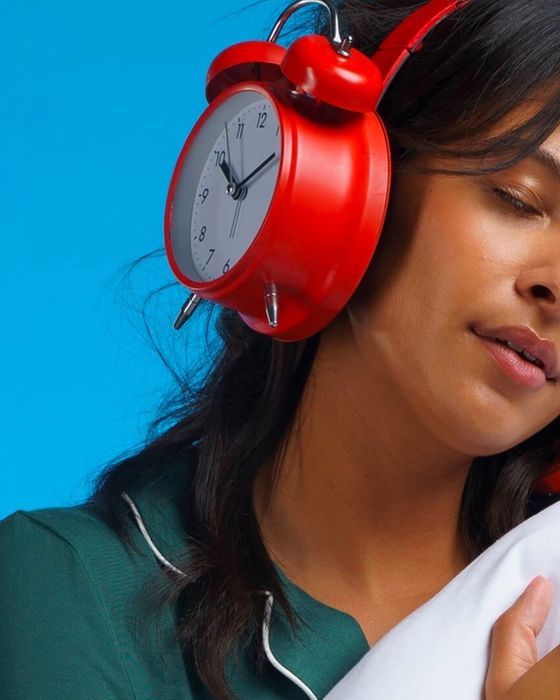 A woman with her eyes closed holding a pillow. Her over-ear headphones are shaped like analog alarm clocks.