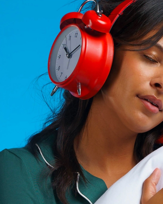 A woman with her eyes closed holding a pillow. Her over-ear headphones are shaped like analog alarm clocks.