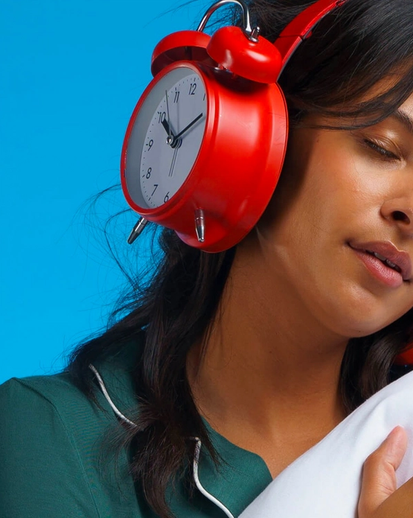 A woman with her eyes closed holding a pillow. Her over-ear headphones are shaped like analog alarm clocks.