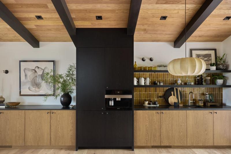 A modern kitchen featuring warm oak lower cabinets, matte black upper cabinetry and beam accents, a golden ochre tile backsplash, open shelving, and a wood-paneled ceiling.