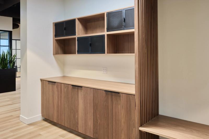 A custom built-in walnut storage unit featuring fluted lower cabinet doors with black bar pulls, an open upper shelving section with perforated black metal mesh cabinet doors, and a vertical slatted wood panel accent, set in a modern office environment.