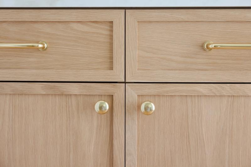 Close-up of light white oak shaker-style cabinet drawers and doors with polished brass bar pulls and round knobs.