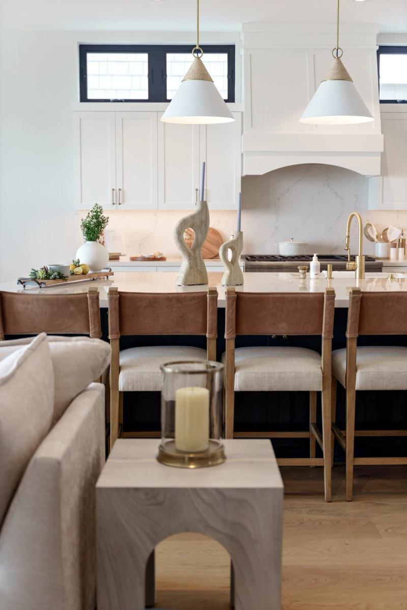 A bright, transitional-style kitchen featuring white upper cabinets, a dark navy kitchen island with a marble countertop, leather and wood bar stools, white cone pendant lights with brass accents, and a marble backsplash behind the range. The foreground shows a living area with a linen sofa and wooden side table.