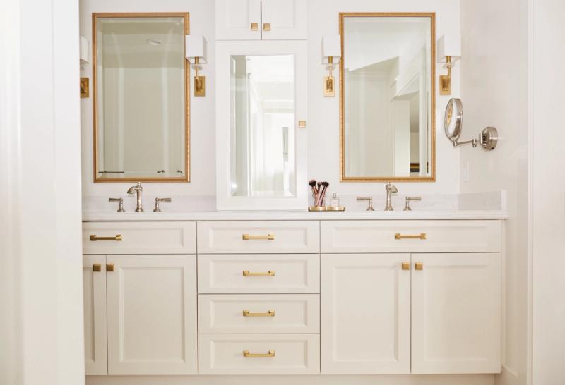 A bright, white double bathroom vanity with raised-panel cabinet doors, gold hardware, two gold-framed mirrors, and wall sconces flanking a center medicine cabinet.