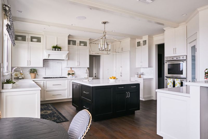 A bright, spacious kitchen featuring white shaker cabinets with chrome hardware, a dark navy-black center island with white quartz countertop, hardwood floors, and a geometric chrome pendant light overhead.