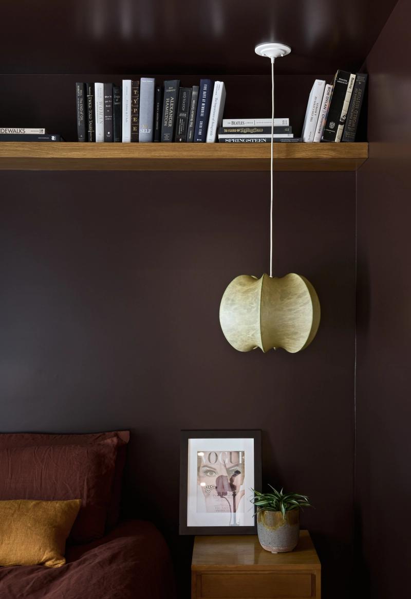 A deep brown bedroom wall featuring a floating oak bookshelf filled with books, a gold cocoon pendant light hanging from the ceiling, and a wood nightstand with a framed Vogue print and small potted plant.