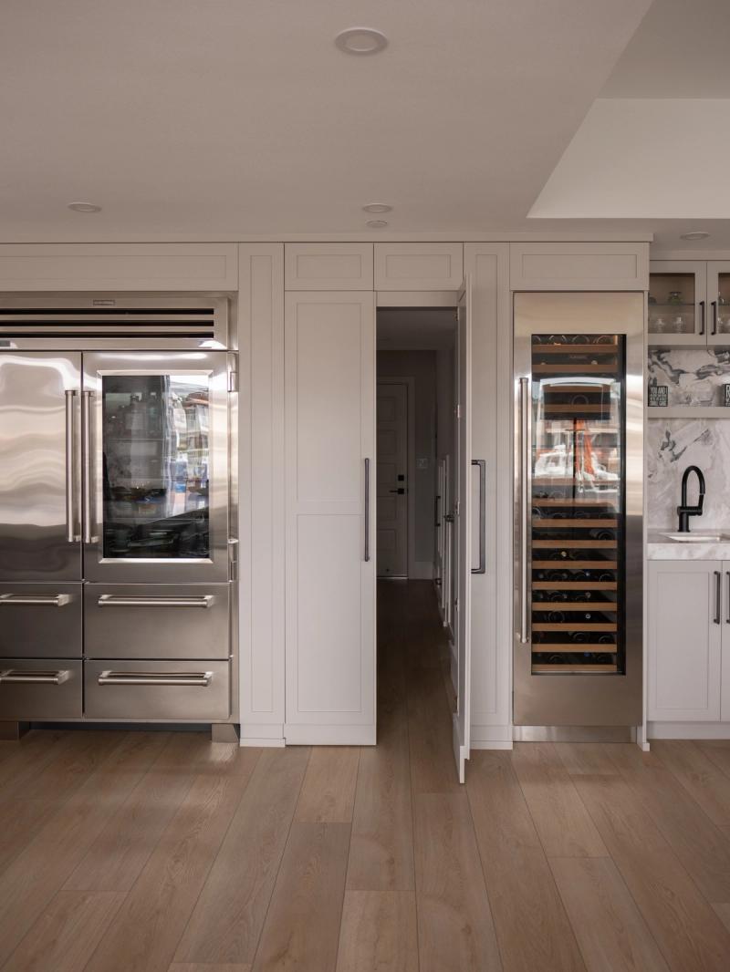 A high-end transitional kitchen featuring floor-to-ceiling white shaker cabinetry, a stainless steel Sub-Zero French door refrigerator, a built-in wine column, and a hidden pantry door, all set on wide-plank hardwood floors.