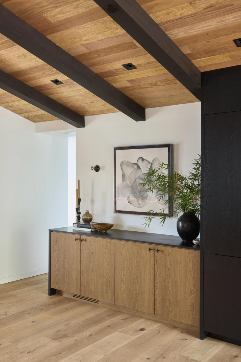 A custom wood credenza with natural oak cabinet doors and a dark matte top, styled with a black vase, bamboo plant, and artwork, set beneath an exposed beam wood-plank ceiling.