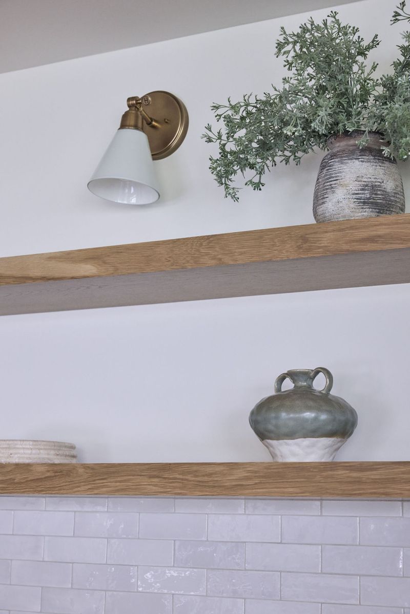 Two natural oak floating shelves mounted on a white wall above subway tile, styled with ceramic vases and a brass wall sconce with a white shade.