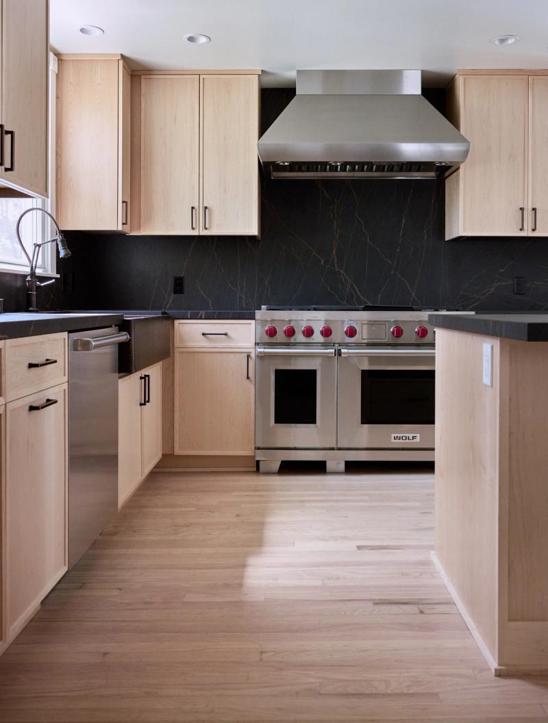 A modern kitchen featuring light maple flat-panel cabinetry, dark soapstone countertops and backsplash, a stainless steel Wolf dual-fuel range with red knobs, and a matching range hood, set against light hardwood floors.