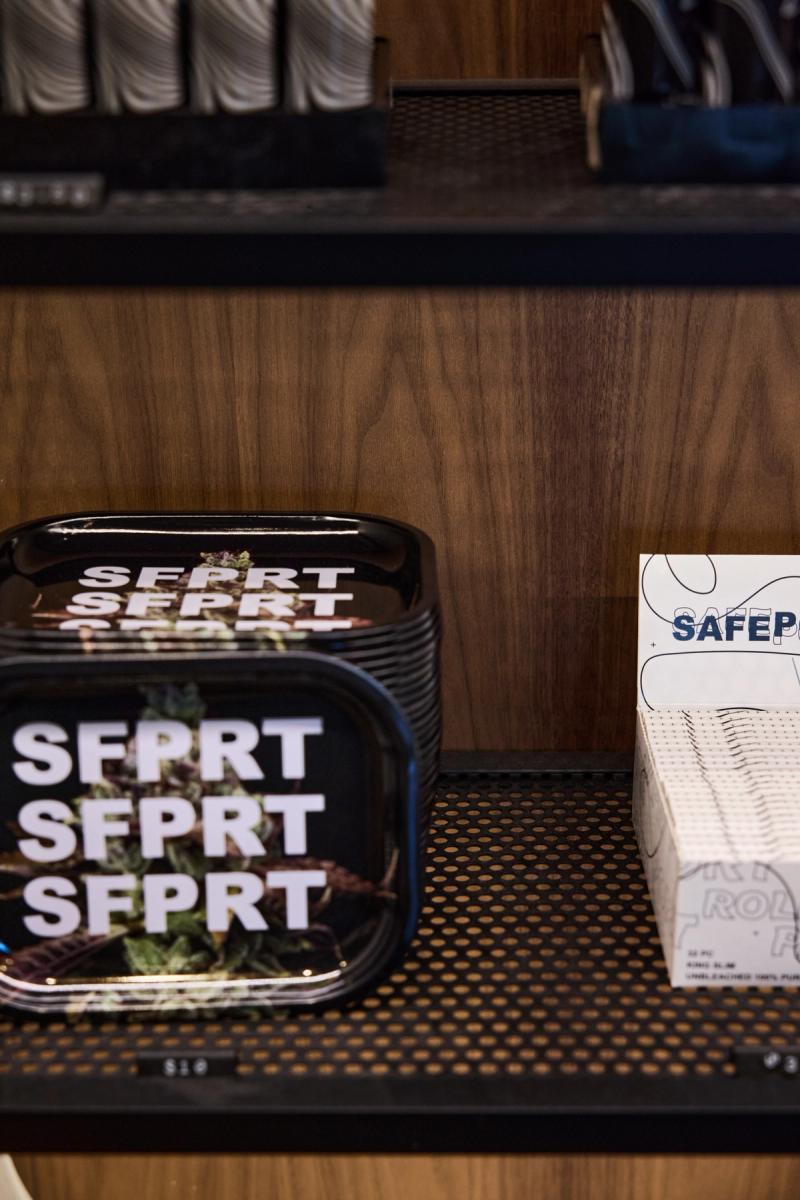A close-up shot of branded SFPRT rolling trays stacked on a perforated black shelf surface, with a box of rolling papers visible to the right, set against a warm walnut wood panel background.