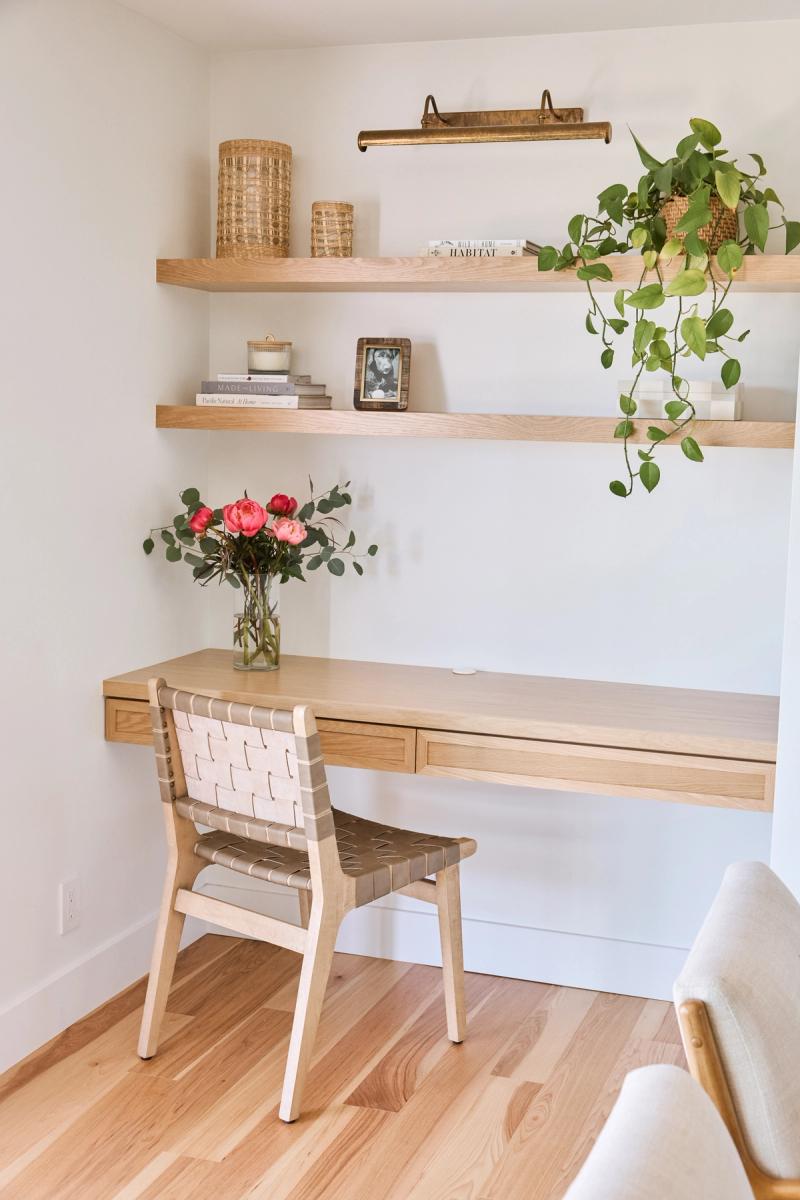 A light wood floating desk with drawers is mounted on a white wall, paired with a woven leather chair and two floating oak shelves styled with books, baskets, a trailing plant, and fresh pink flowers.